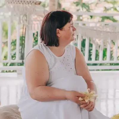 A woman dressed in white sitting on the floor, holding a flower, during a Balinese ceremony outdoors with trees and plants in the background.