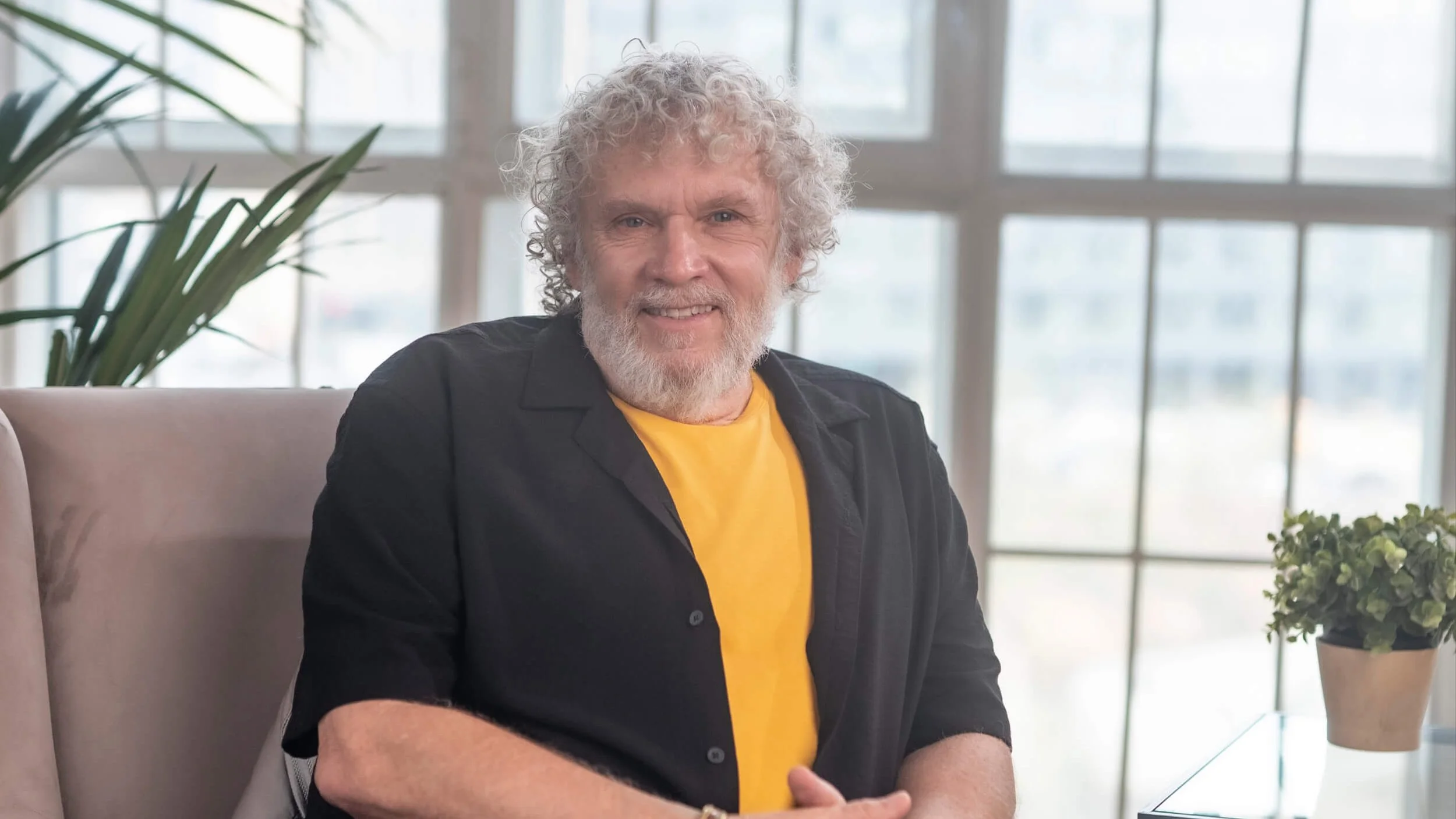 A smiling older man with curly gray hair and a beard sitting indoors in front of large windows, wearing a black shirt over a yellow T-shirt, with plants in the background.