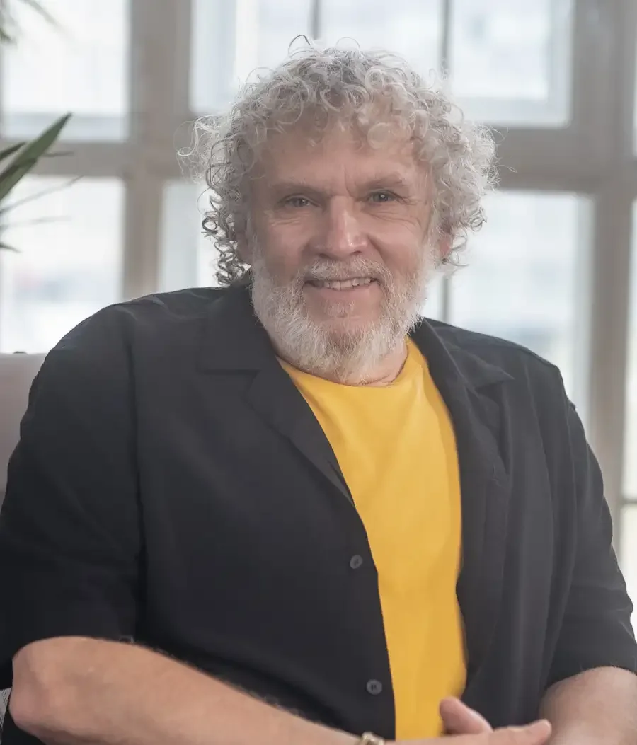 A smiling older man with curly gray hair and a beard sitting indoors in front of large windows, wearing a black shirt over a yellow T-shirt, with plants in the background.