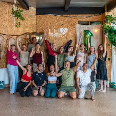 A group of people celebrating indoors with palms and peace signs, in front of a wooden wall decorated with dreamcatchers, greenery, and balloons, including a large gold heart and a green number 1 balloon, with a sign that reads 'Luv Yourself'.
