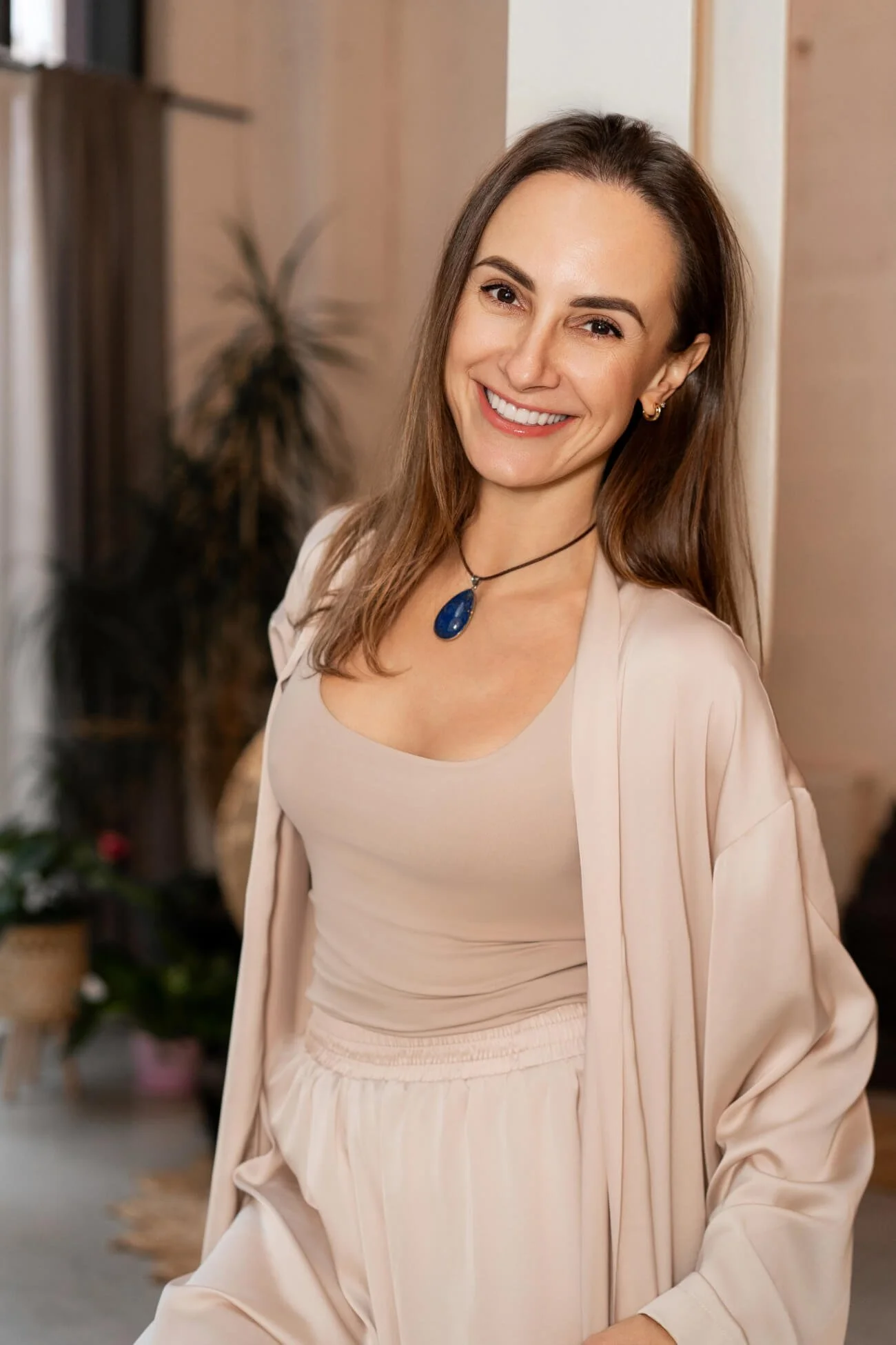 A smiling woman with brown hair wearing a beige outfit and a blue pendant necklace, standing indoors with a blurred background.