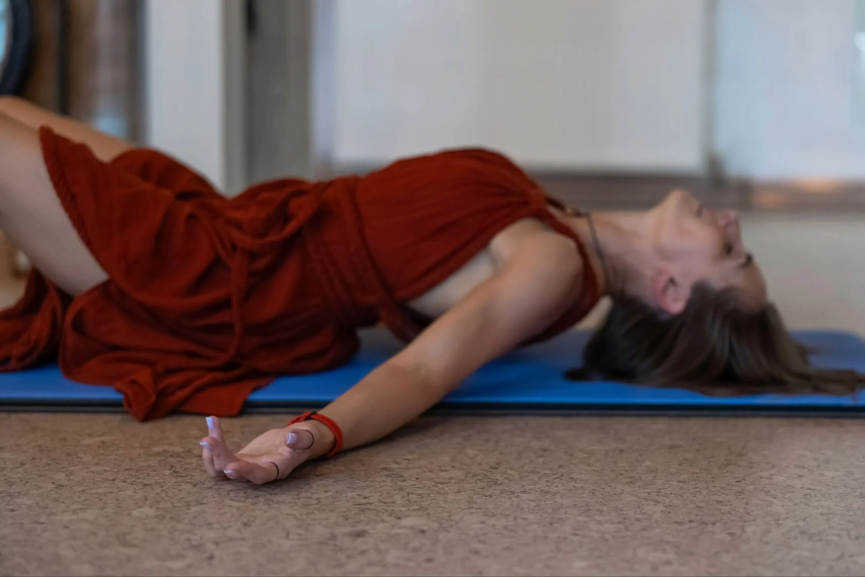 A woman with long brown hair in a red dress lying on a blue yoga mat on the floor, stretching with one arm extended forward and face turned upward.