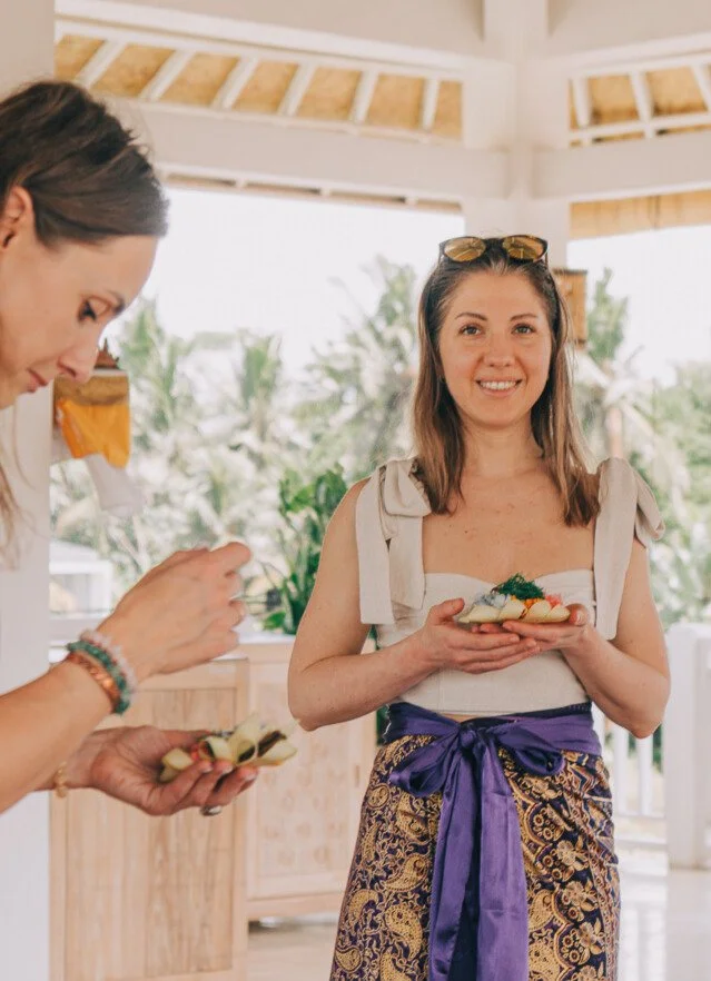 A woman smiling and holding a plate of food, standing outdoors in a tropical setting, with palm trees in the background. Wearing Balinese sarong.
