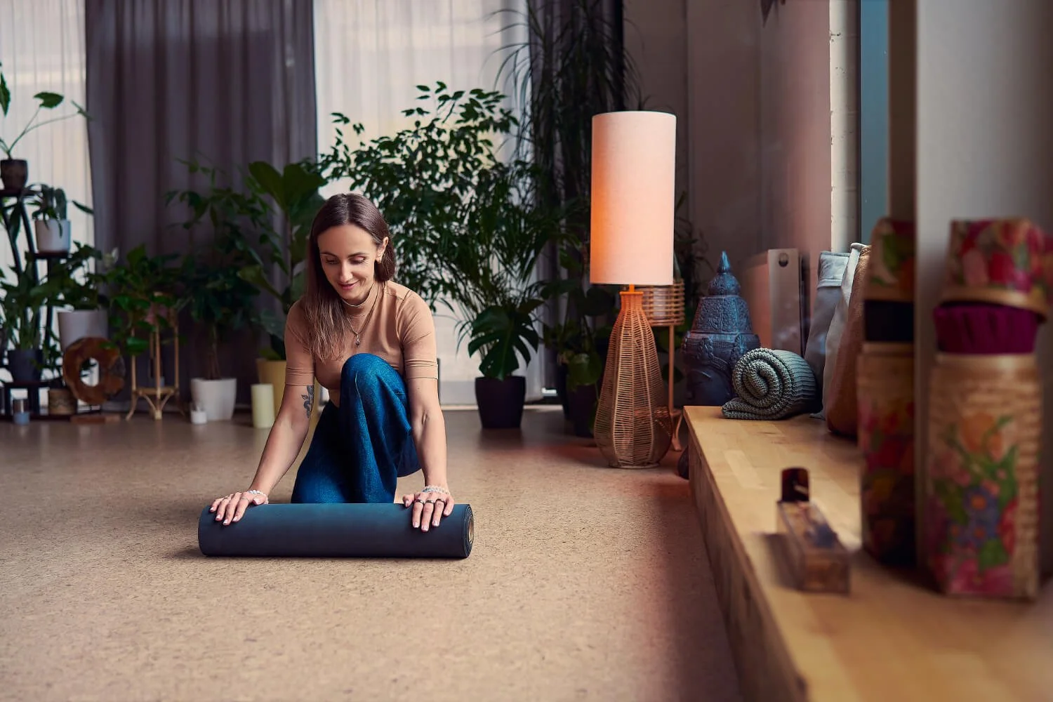Woman in a tan top and blue jeans rolling up a yoga mat in a cosy, artistic room with plants, a tall lamp, and decorative items.
