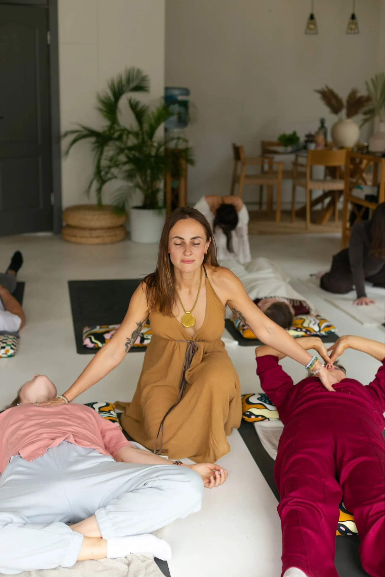 A woman facilitating the energy with her eyes closed while sitting on the floor, surrounded by people lying down or sitting with eyes closed in a cosy, well-lit room.