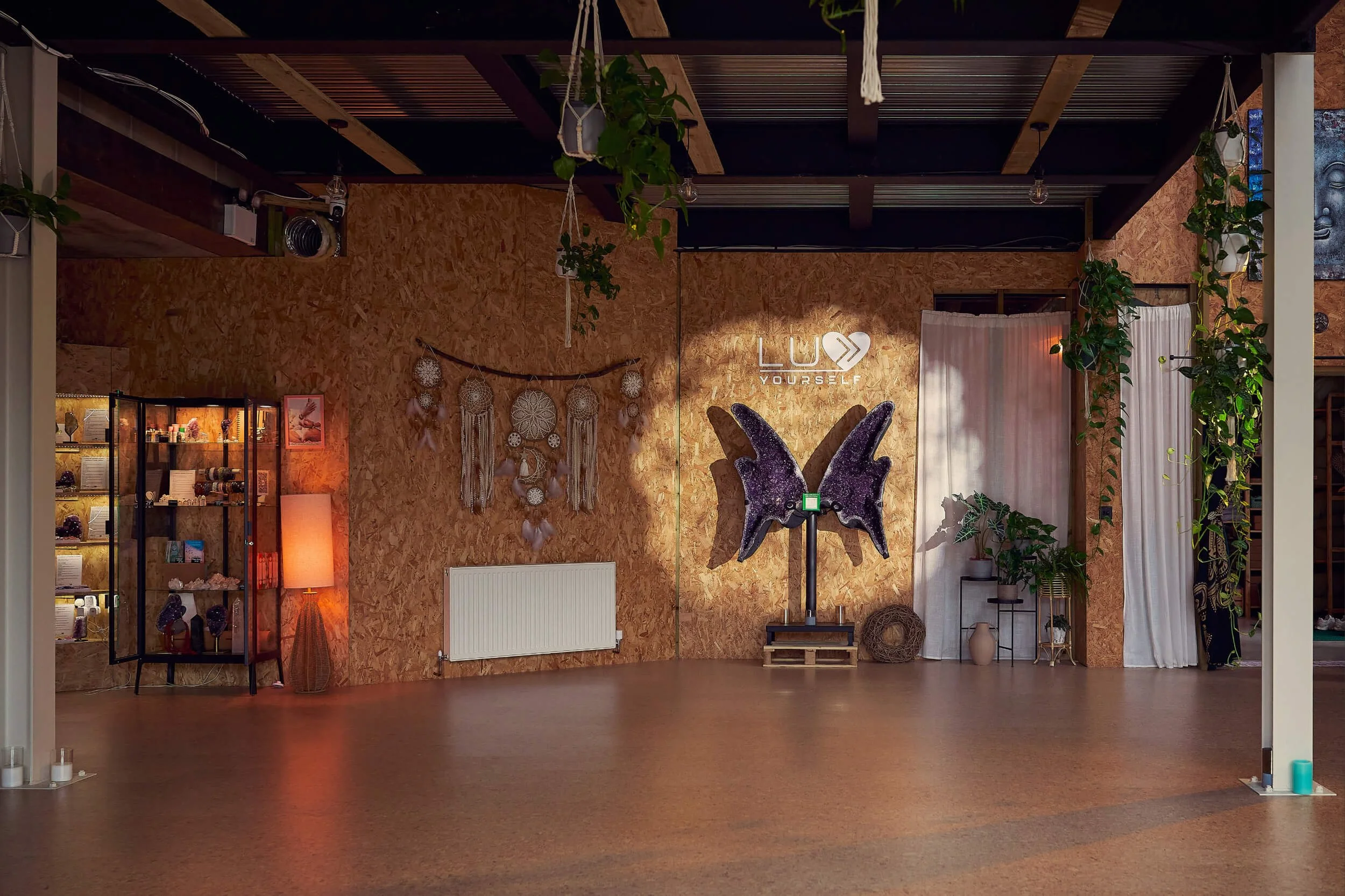 Interior of a yoga or meditation studio with wooden walls, decorative dreamcatchers, large butterfly-shaped amethyst crystal wings, potted plants, and a neon sign that says 'LUV YOURSELF'