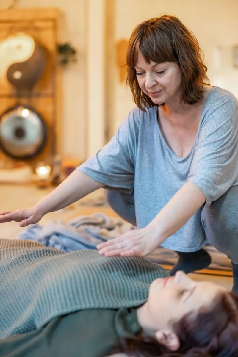 A woman practicing reiki or energy healing on a woman lying down in a yoga pose in a peaceful space.