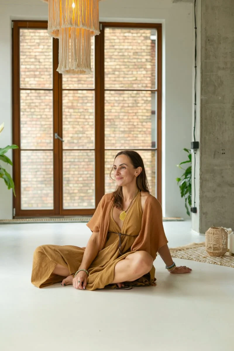 A woman sitting cross-legged on the floor inside a room with large windows and brick exterior visible outside. She is wearing a brown dress and accessorized with jewelry, smiling softly.