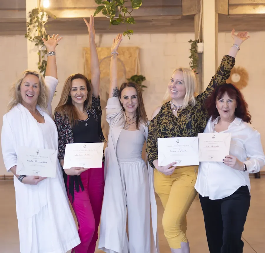 Five women standing together, some holding certificates, celebrating at an indoor event, with their arms raised and smiling. Finishing Kundalini Activation facilitator training with Natalia Bareikis as it says on certificate.