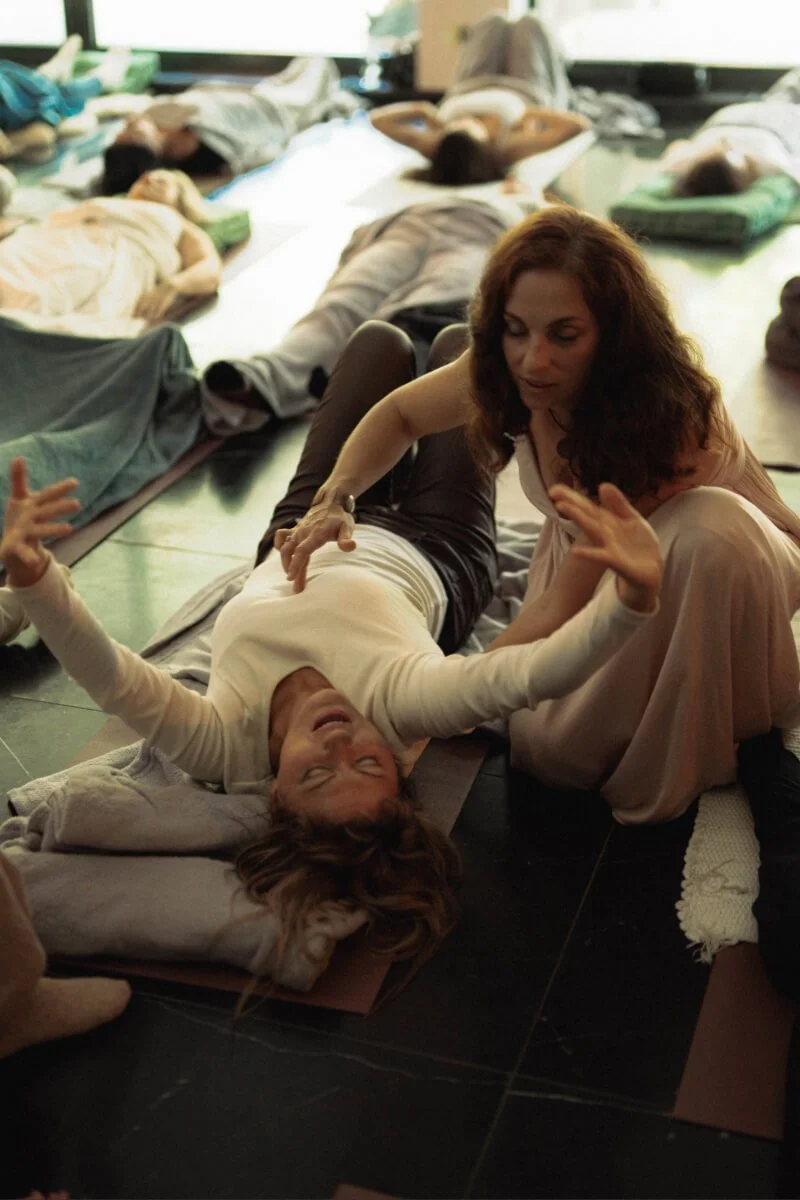 A woman and a girl doing healing session on a mat in a room with several other people lying down on mats behind them.