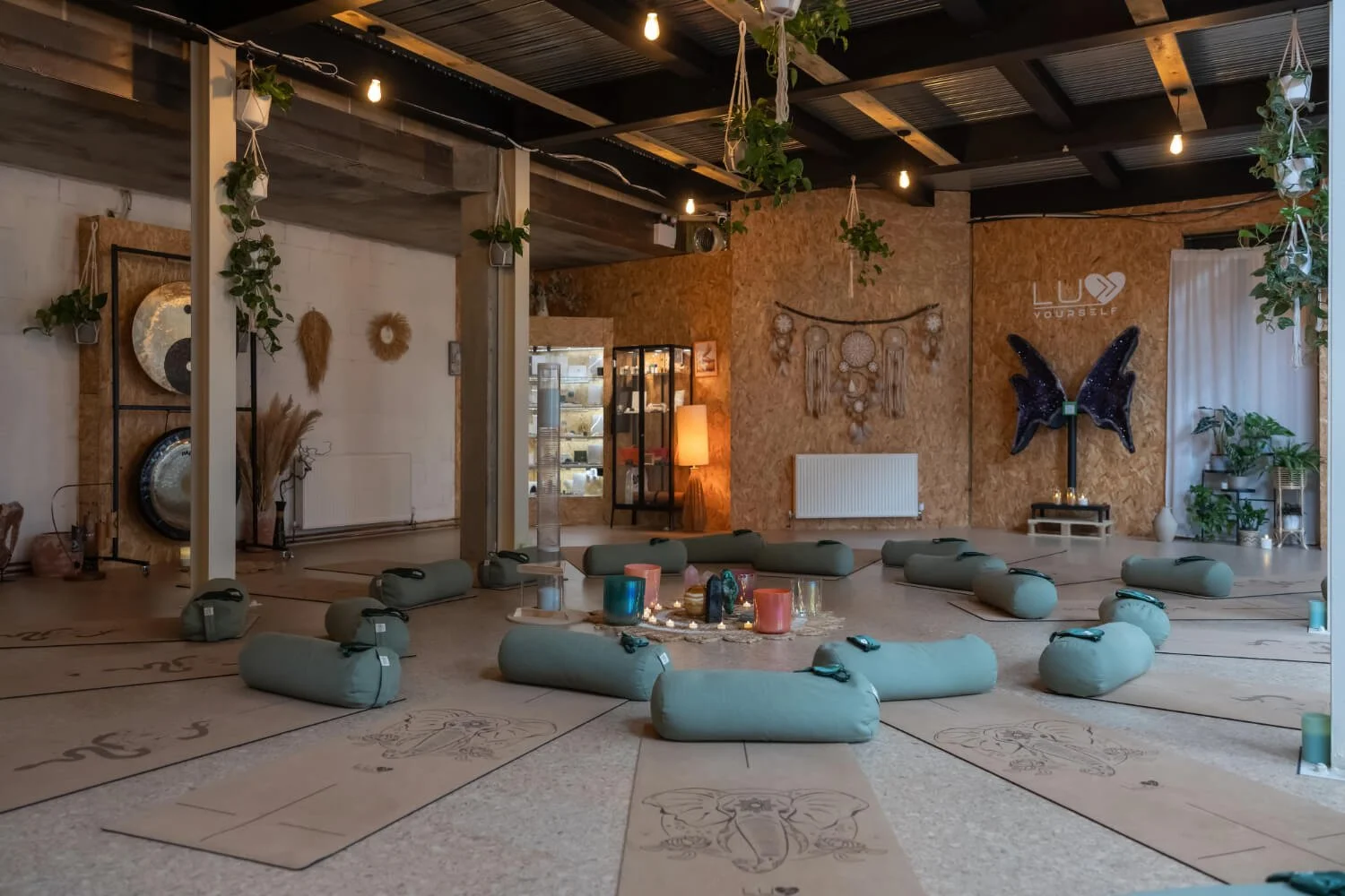 A cosy wellness space with yoga mats featuring kundalini snake and elephant head, bolsters, candles, and hanging plants, decorated with wall art and fairy lights. Wall has a sign LUV YOURSELF and by the side a butterfly amethyst crystal wings.