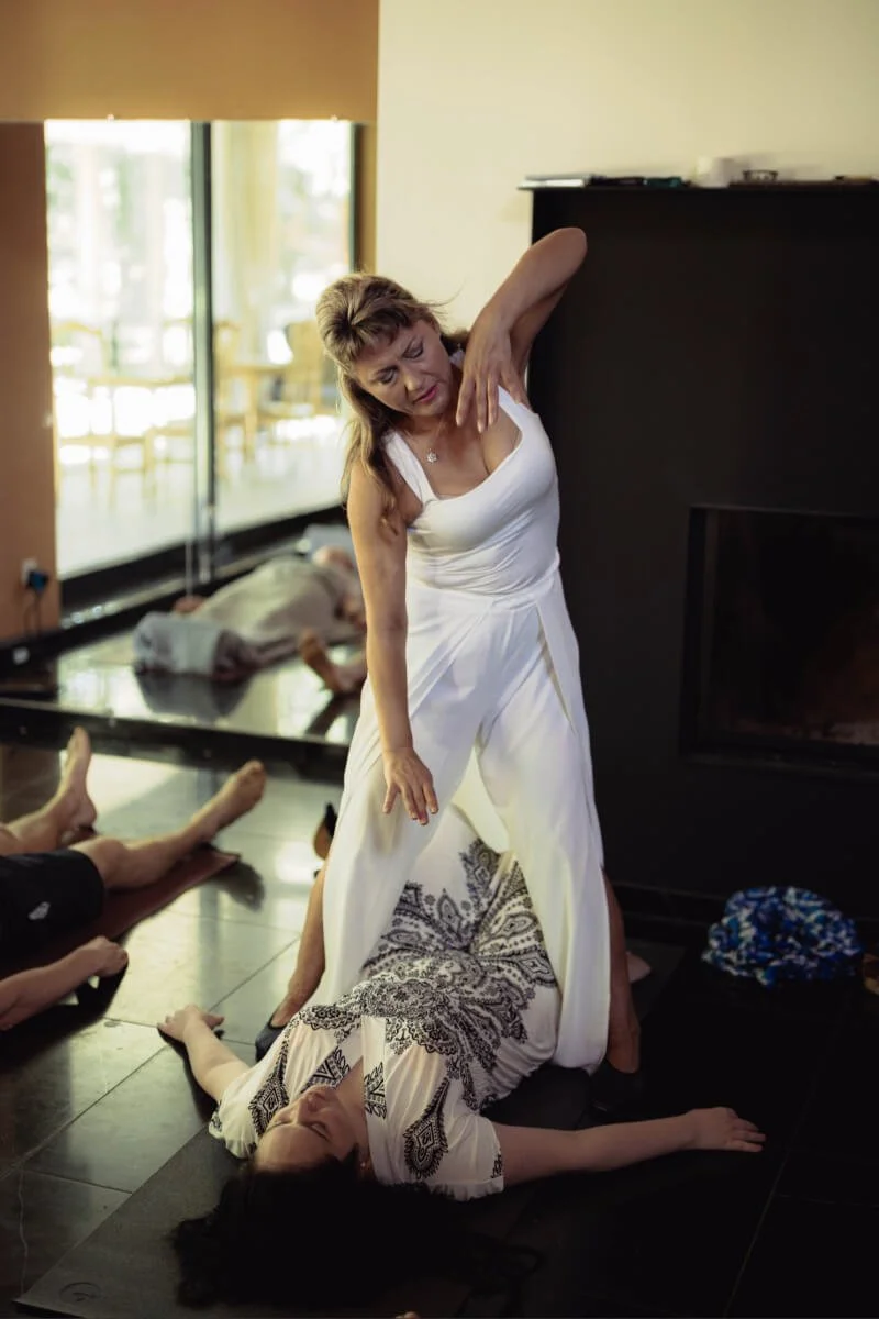 A woman in a white dress performs a healing session on a woman in a patterned dress lying on the floor in a living room with large windows and a fireplace.