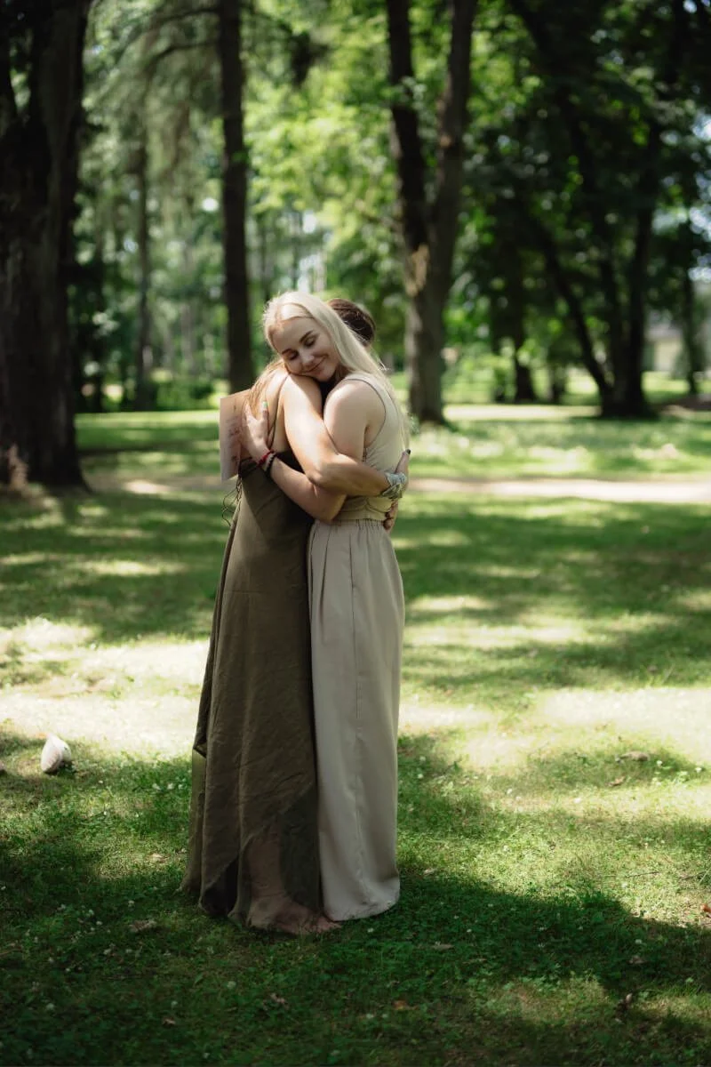 Two women embracing in the nature with tall trees and sunlight filtering through the leaves. They are calm and peaceful.