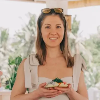 A woman smiling and holding a plate of food, standing outdoors in a tropical setting, with palm trees in the background. Wearing Balinese sarong.