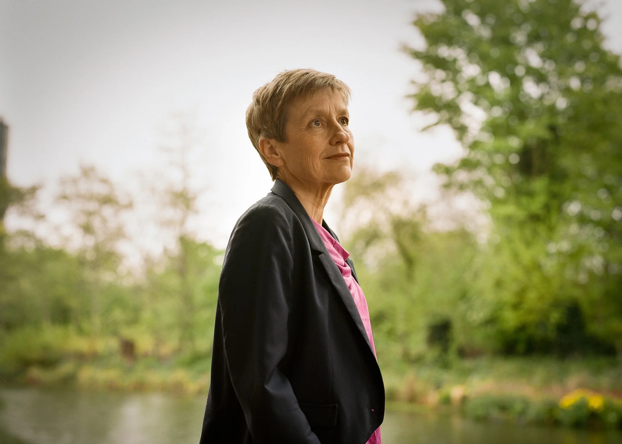 Older woman with short hair, wearing a black blazer over a pink top, standing outdoors in front of a body of water and green trees.