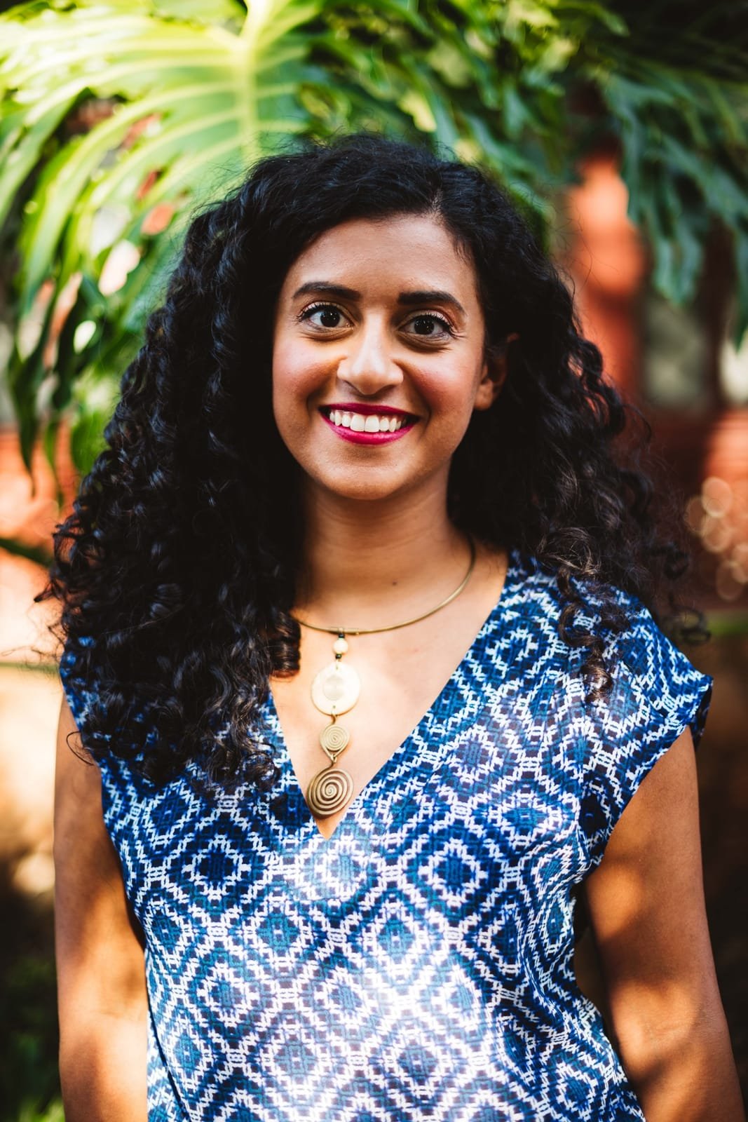 A woman with curly black hair, wearing a blue patterned top and a gold necklace, smiling outdoors in front of green foliage.