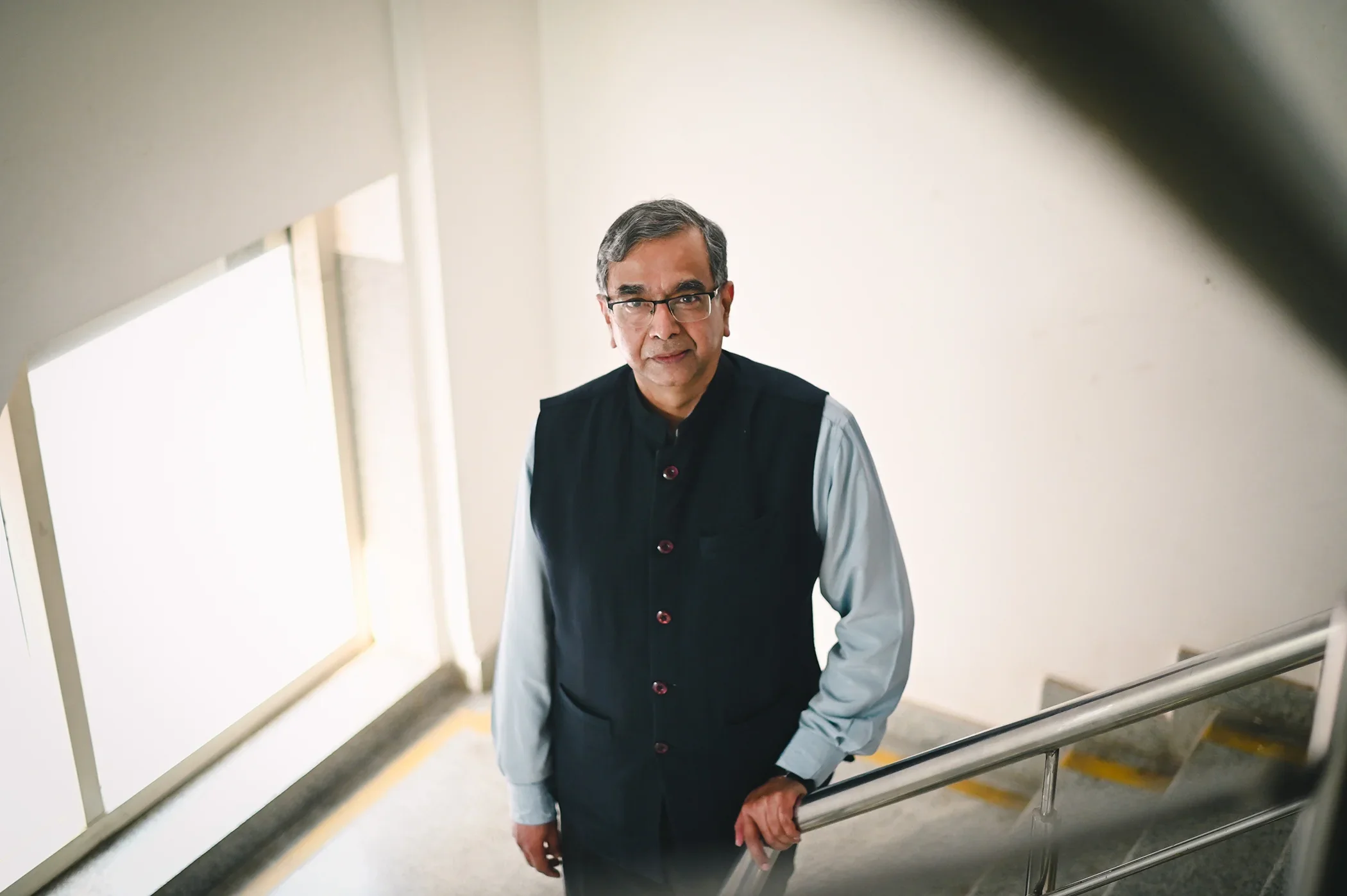 An older man with glasses and grey hair standing on a staircase, wearing a black vest over a light-colored shirt, looking up at the camera.