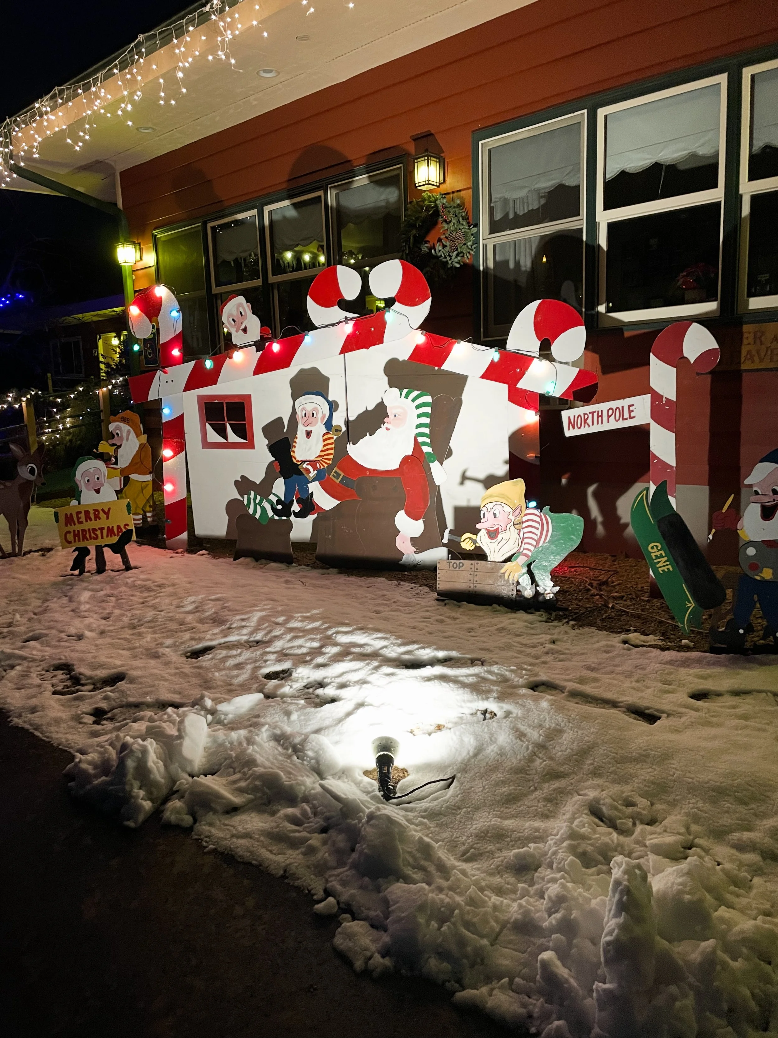 Christmas display outside a house with cartoon Santa, elves, reindeer, and candy cane decorations, brightly lit with Christmas lights and a snow-covered ground.
