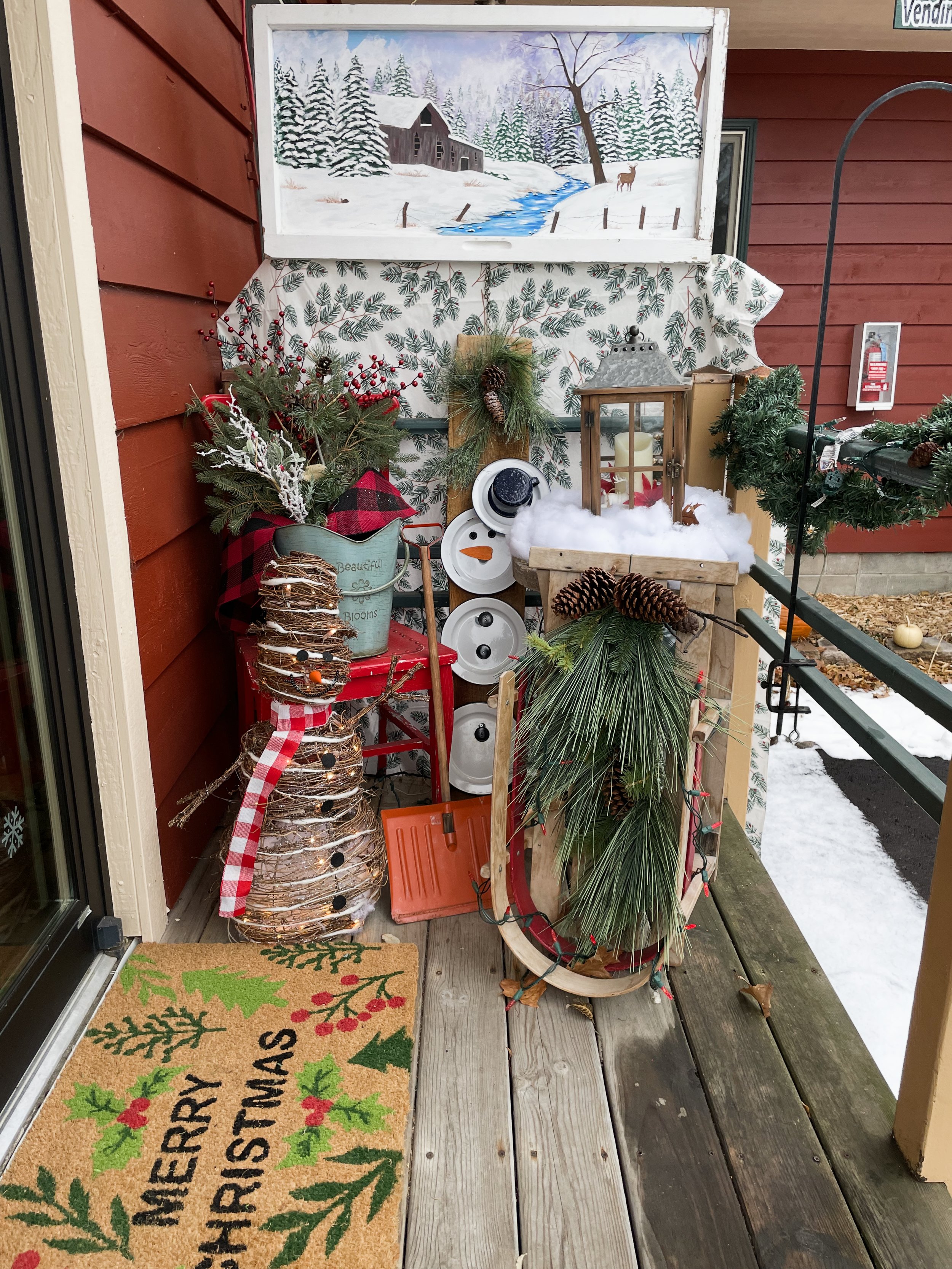 Christmas porch decorated with holiday greenery, a snowman made of twigs with a scarf, a sled with pine branches and pinecones, a lantern surrounded by artificial snow, and a colorful winter-themed doormat that reads 'Merry Christmas'.