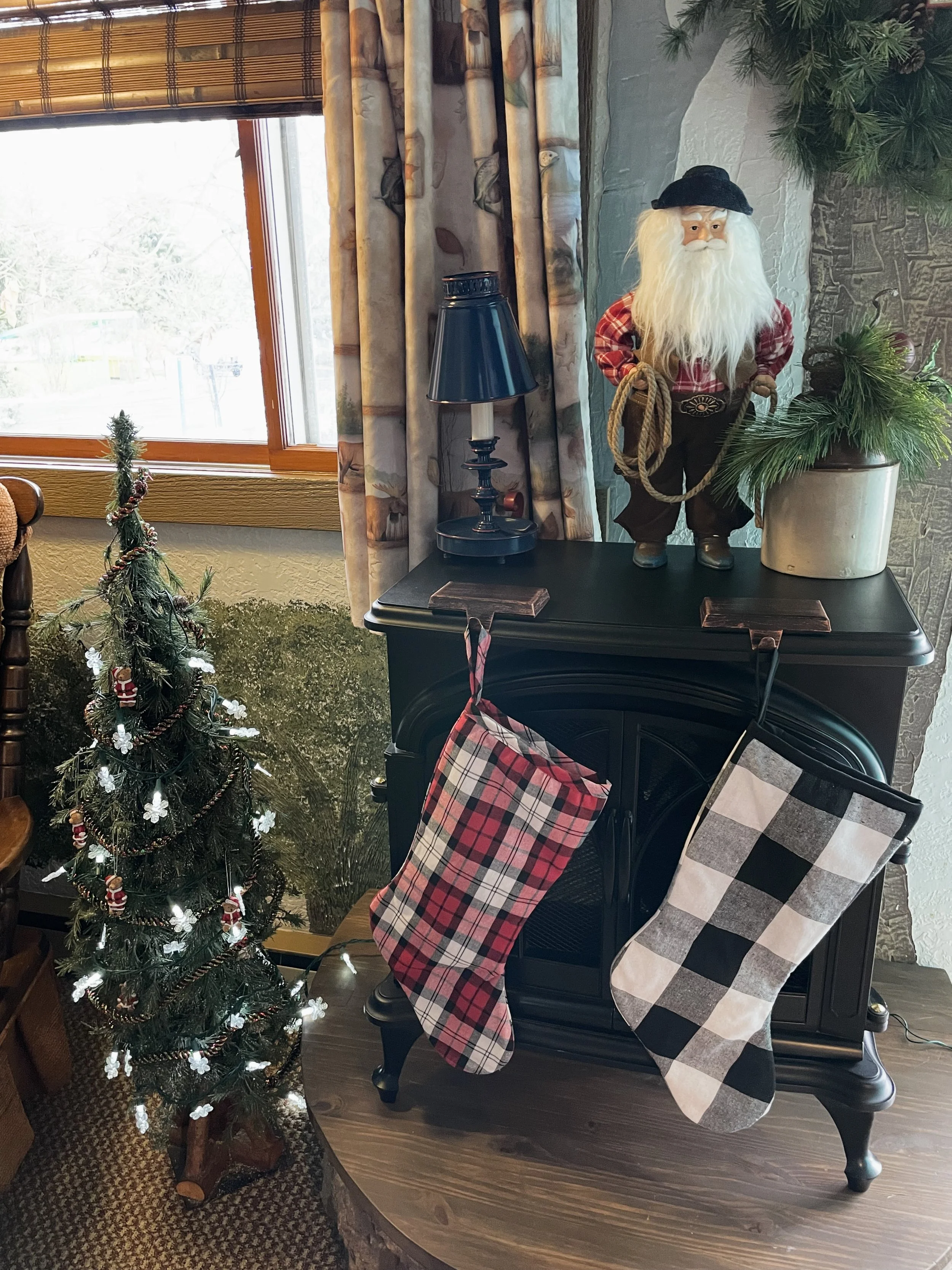 A small decorated Christmas tree with string lights and ornaments is placed next to a black fireplace mantel. On top of the mantel, there is a Santa Claus figure holding a rope, a black lamp, and a potted pine plant. Two Christmas stockings hang from