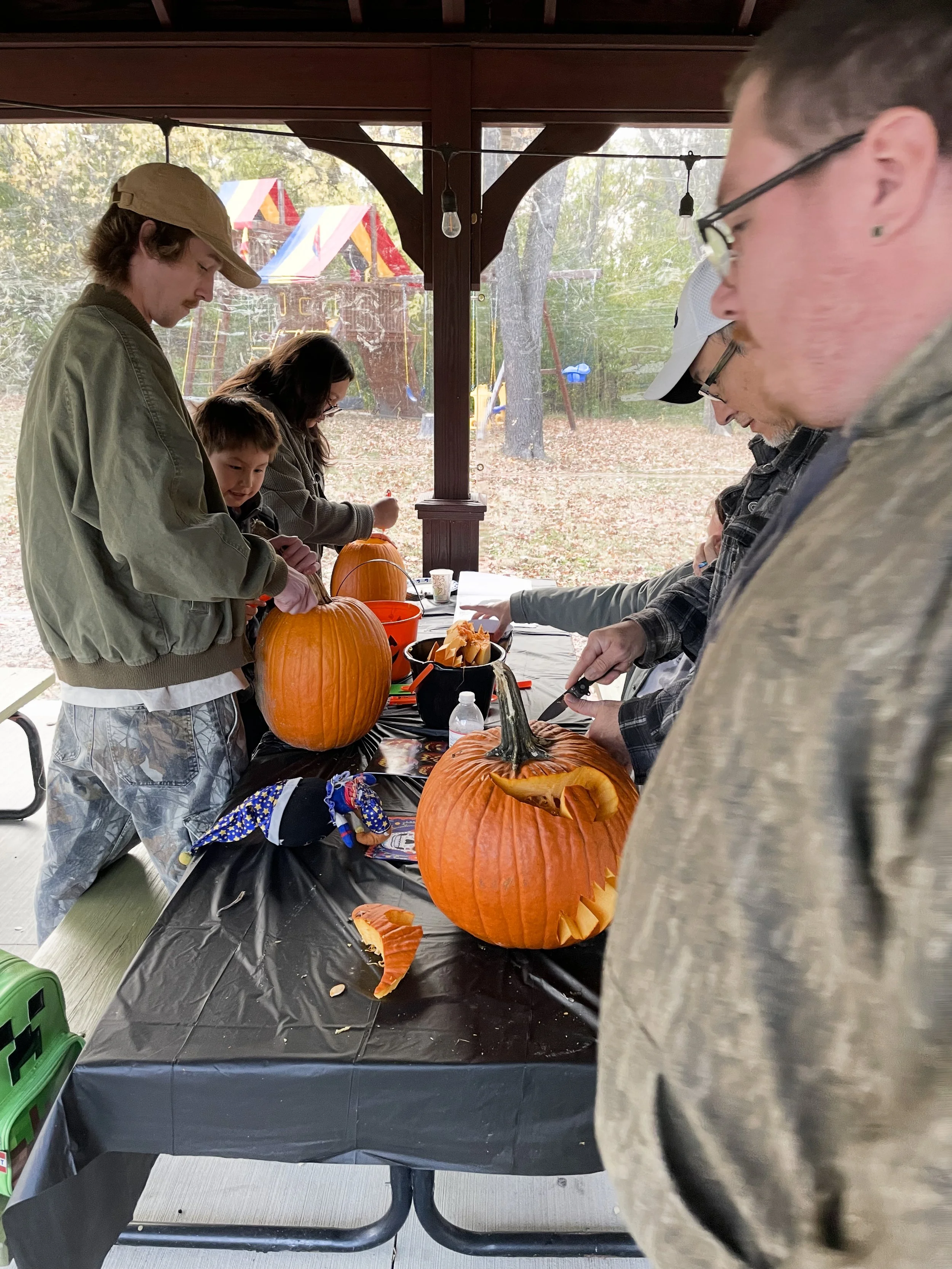 People carving pumpkins at a fall festival under a pavilion, with a playground visible outside.