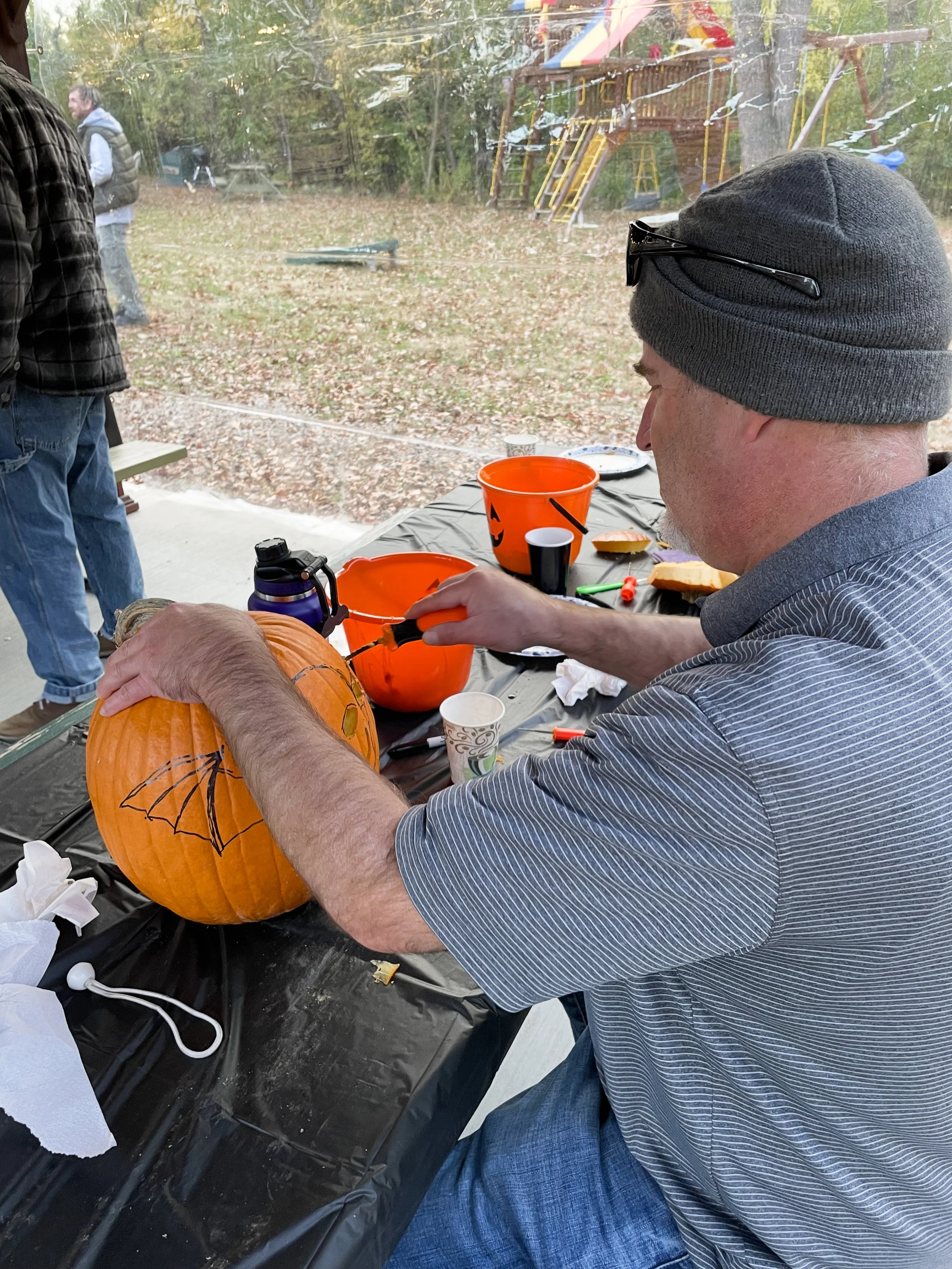 Man carving a bat wing design on a pumpkin at a Halloween party outdoors, with fall leaves and a playground in the background.
