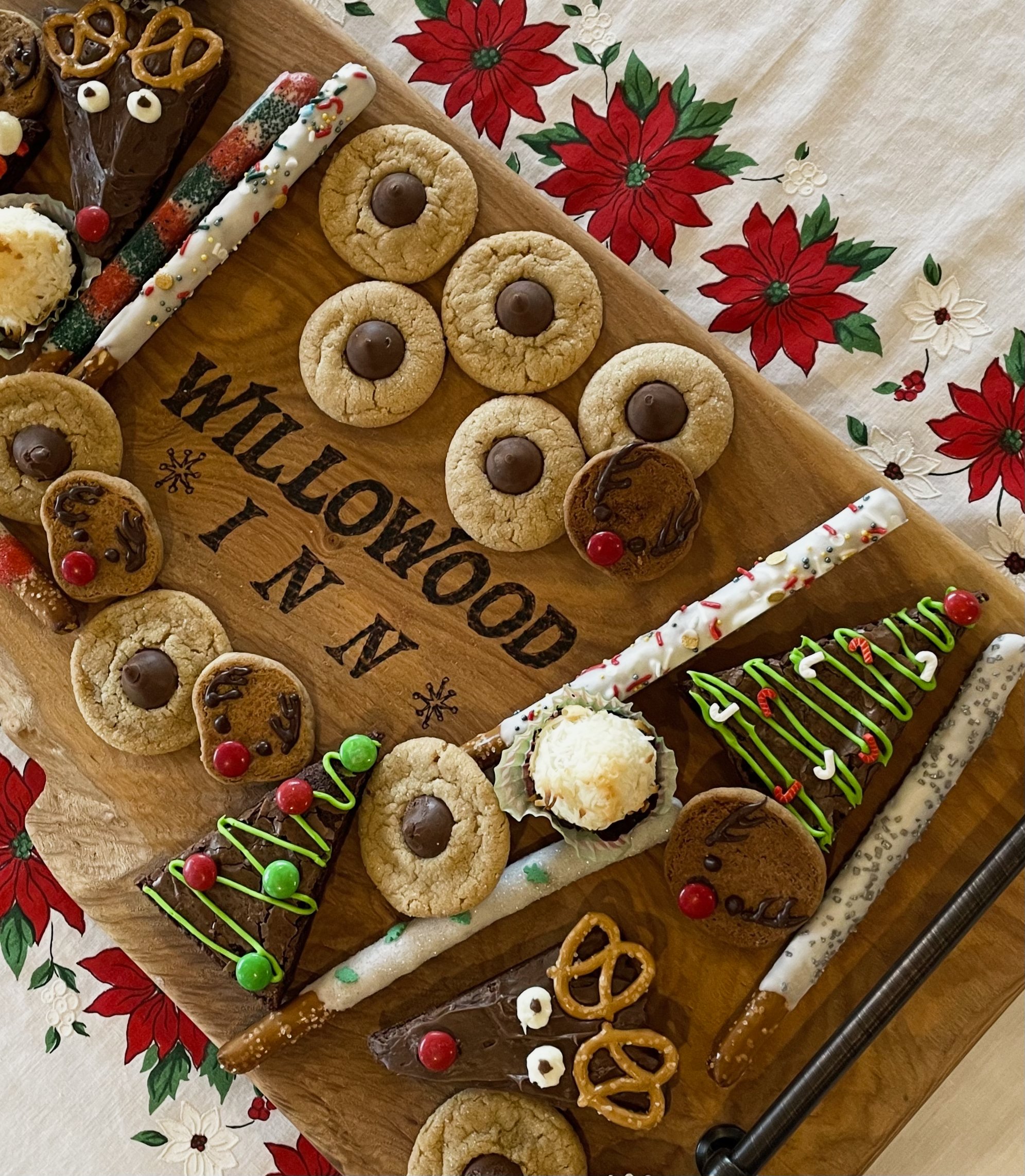 A wooden board with various Christmas-themed cookies and pretzels, decorated with green and red icing, candies, and a white chocolate pretzel. The board has a message written on it that says 'MERRY CHRISTMAS' and some text that is reversed.