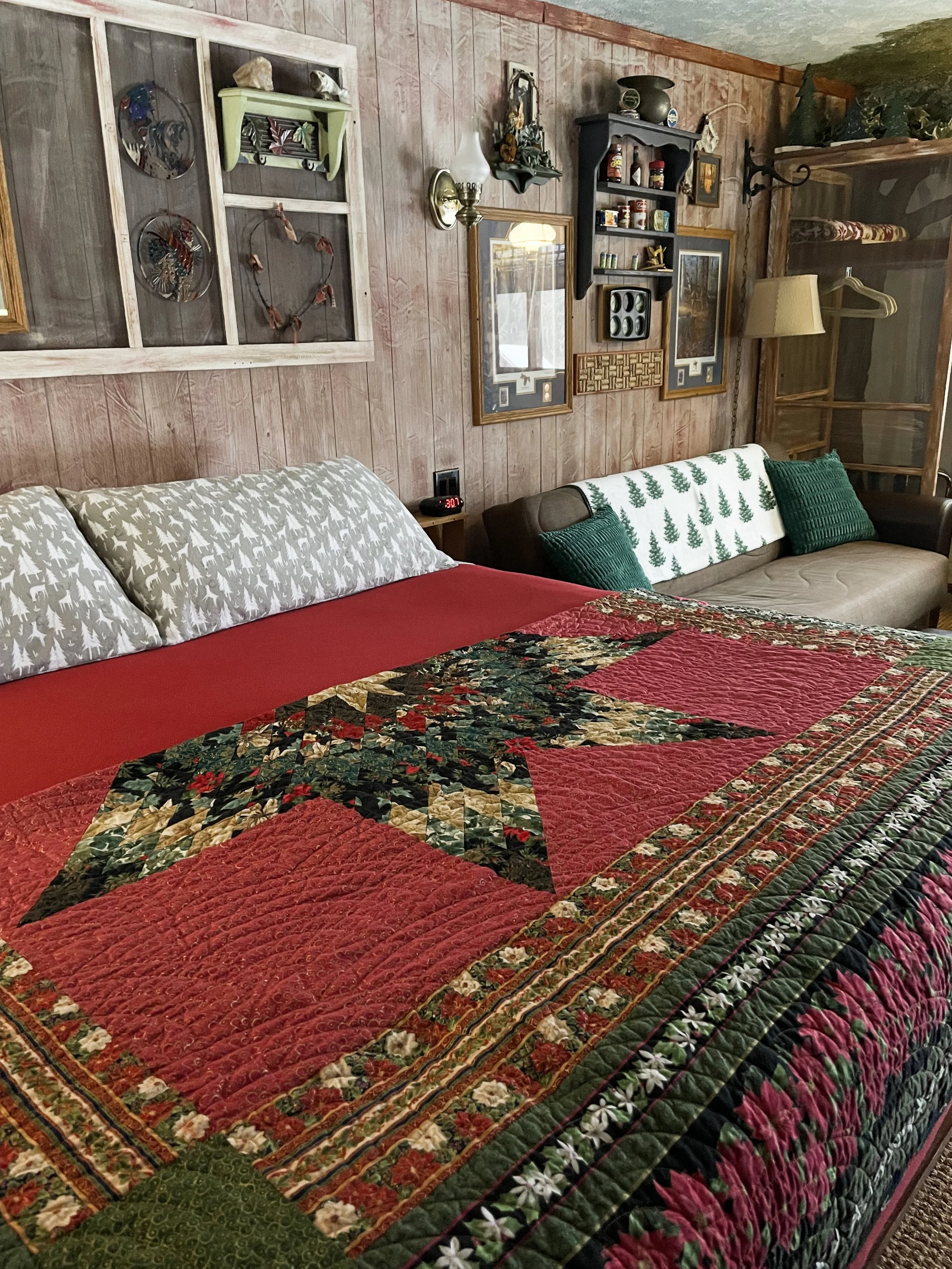 A cozy bedroom with wood-paneled walls, featuring a bed with a festive Christmas quilt, pillows with forest and tree patterns, and a vintage aesthetic with various wall decorations, including framed pictures, shelves, and decorative plates.