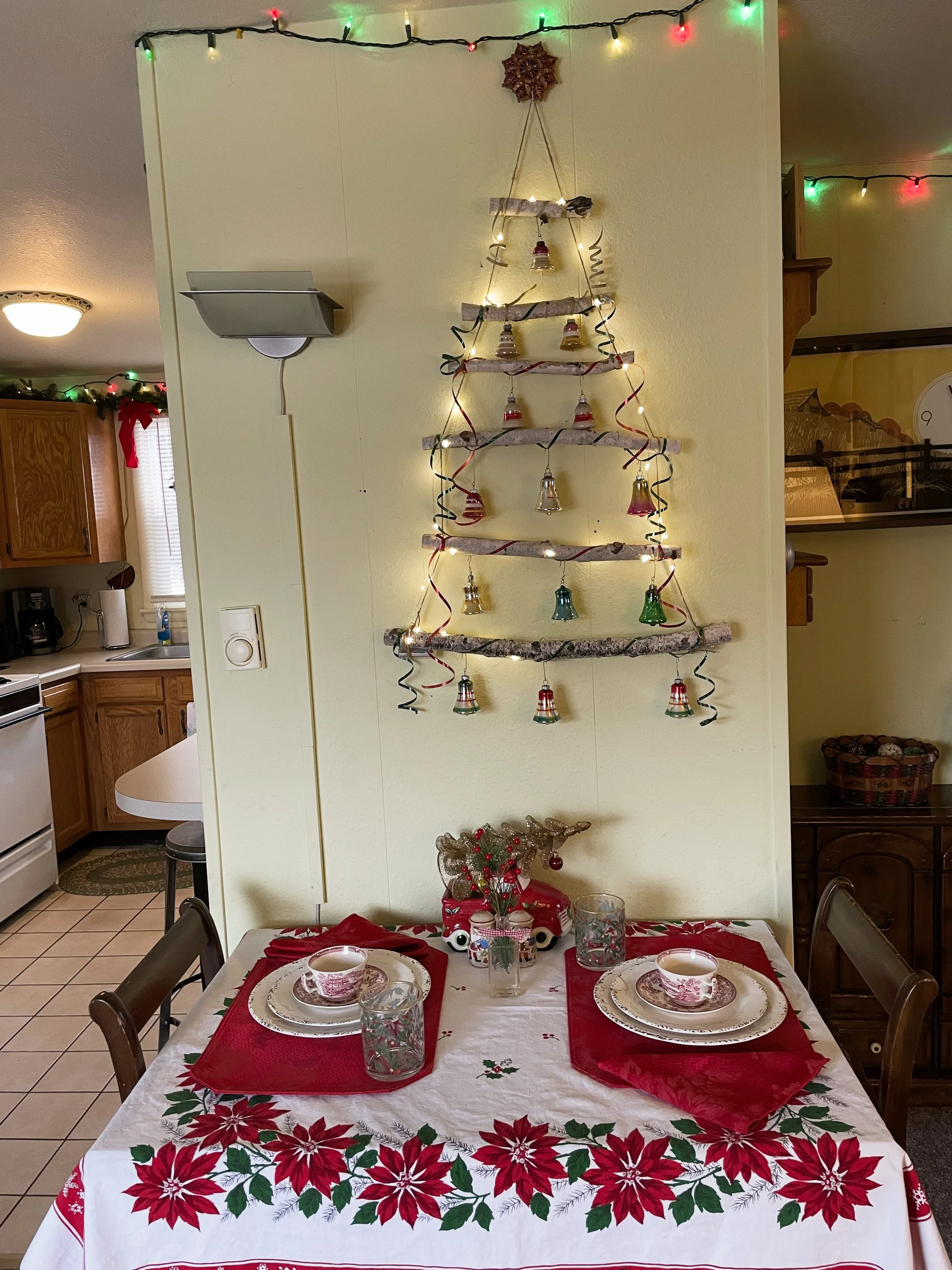 A dining table decorated with a Christmas tablecloth, place settings, and holiday decorations. A wall hanging made of driftwood is arranged in a triangular shape to resemble a Christmas tree, decorated with small bells and curling ribbons. String lig