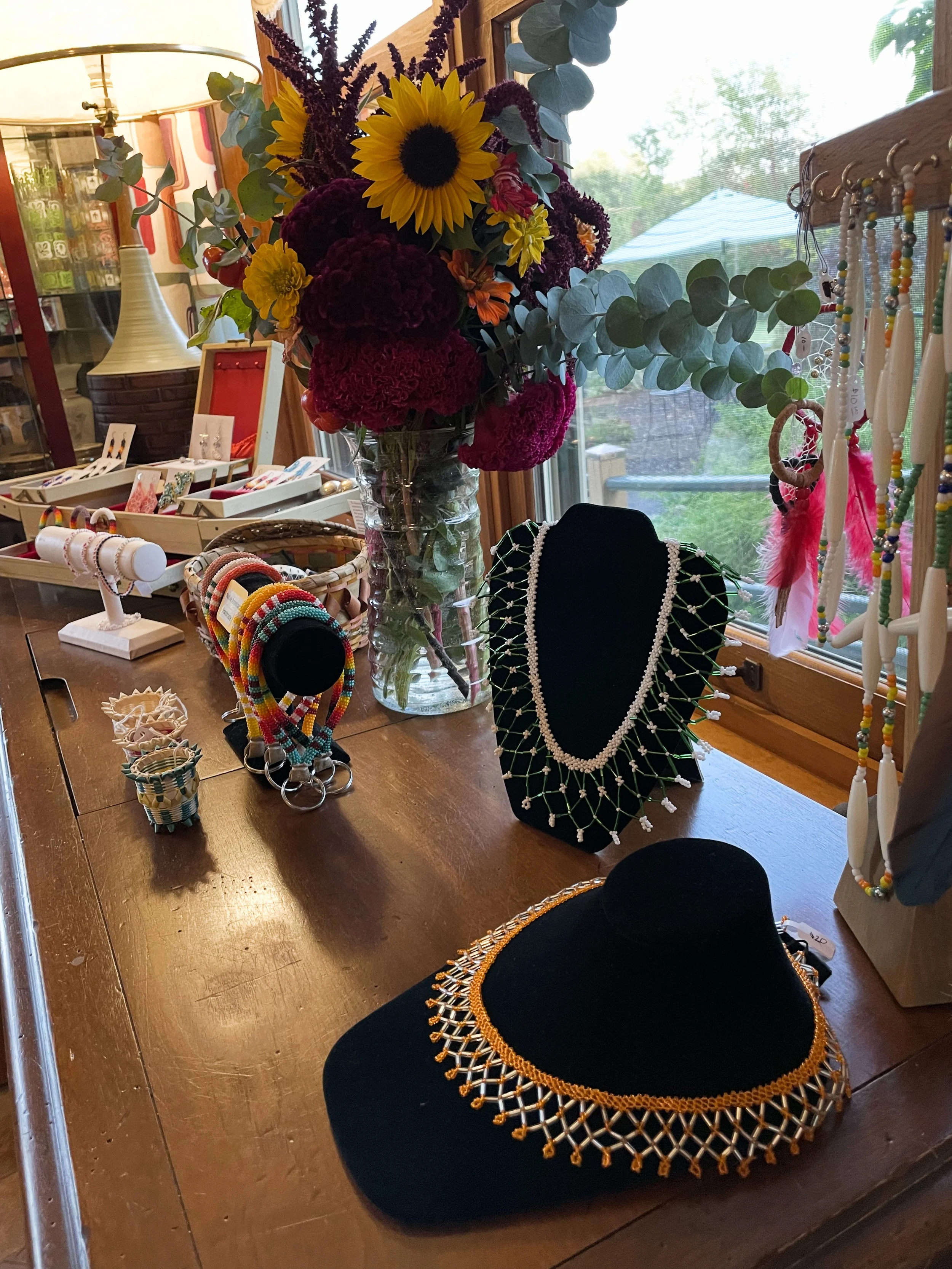Jewelry display with necklaces, bracelets, and earrings, alongside a large bouquet of sunflowers and other colorful flowers on a table near a window.