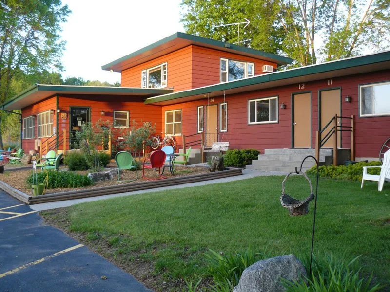 A modern red building with multiple levels and large windows, surrounded by a well-maintained garden with colorful chairs and greenery.
