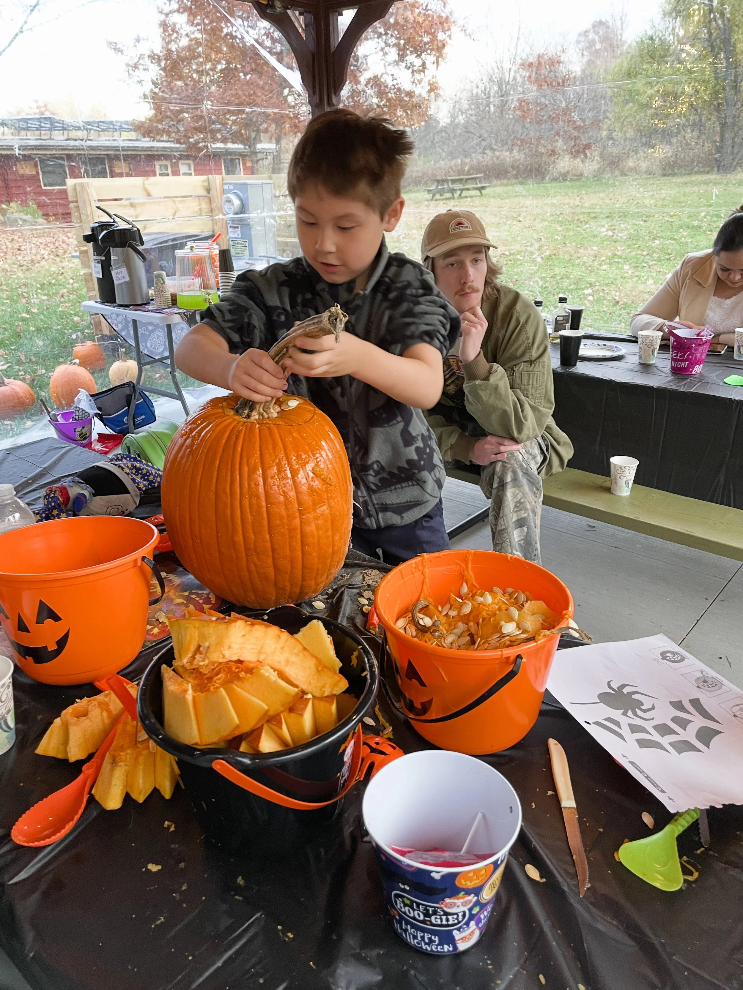 A young boy carving a pumpkin at a Halloween-themed party, surrounded by pumpkin-shaped buckets, pumpkin slices, and Halloween decorations, with autumn trees and pumpkins outside the window.