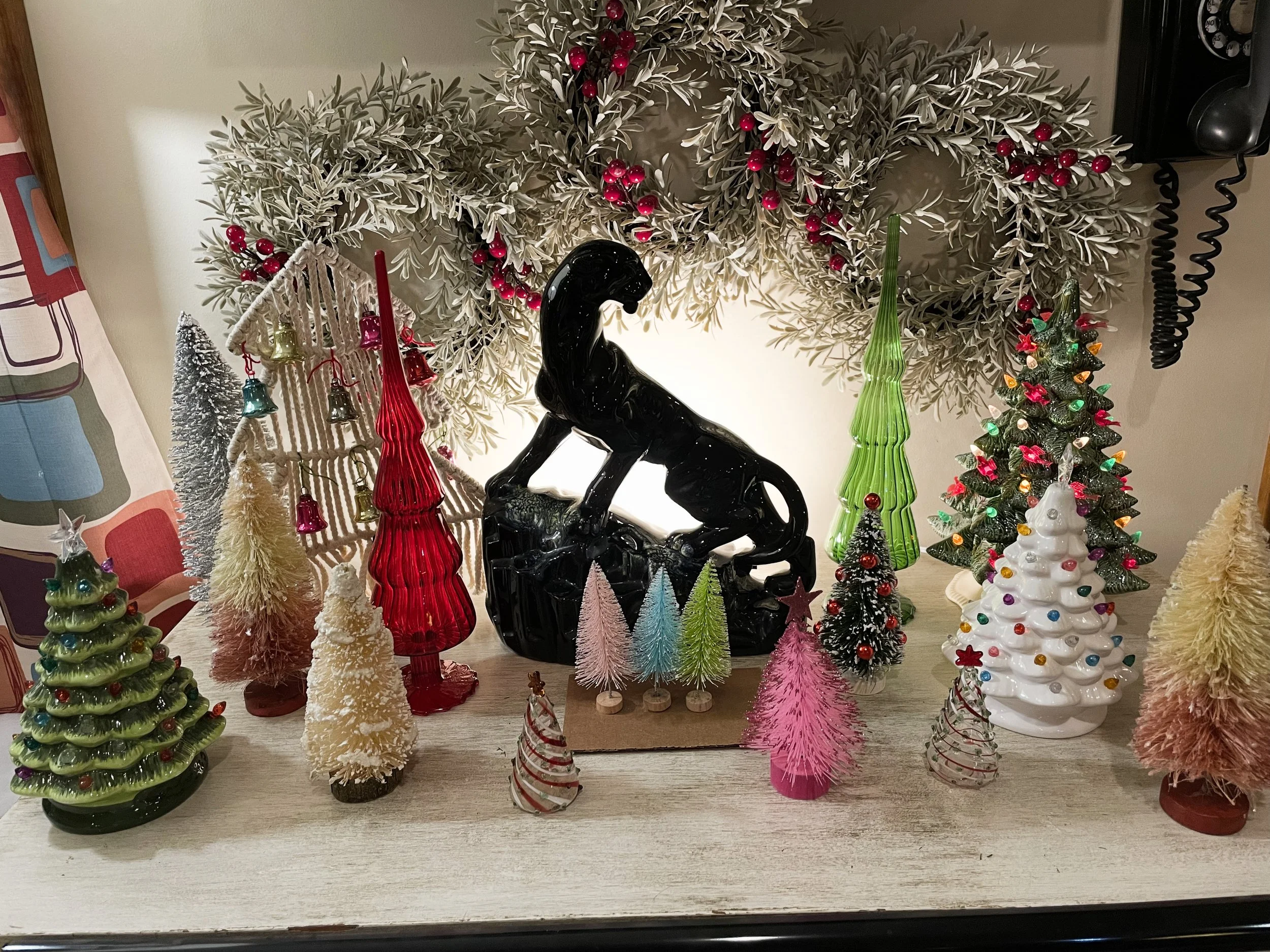 Decorative Christmas display with various festive glass Christmas trees, a black horse figurine, and a garland of frosted leaves with red berries in the background.