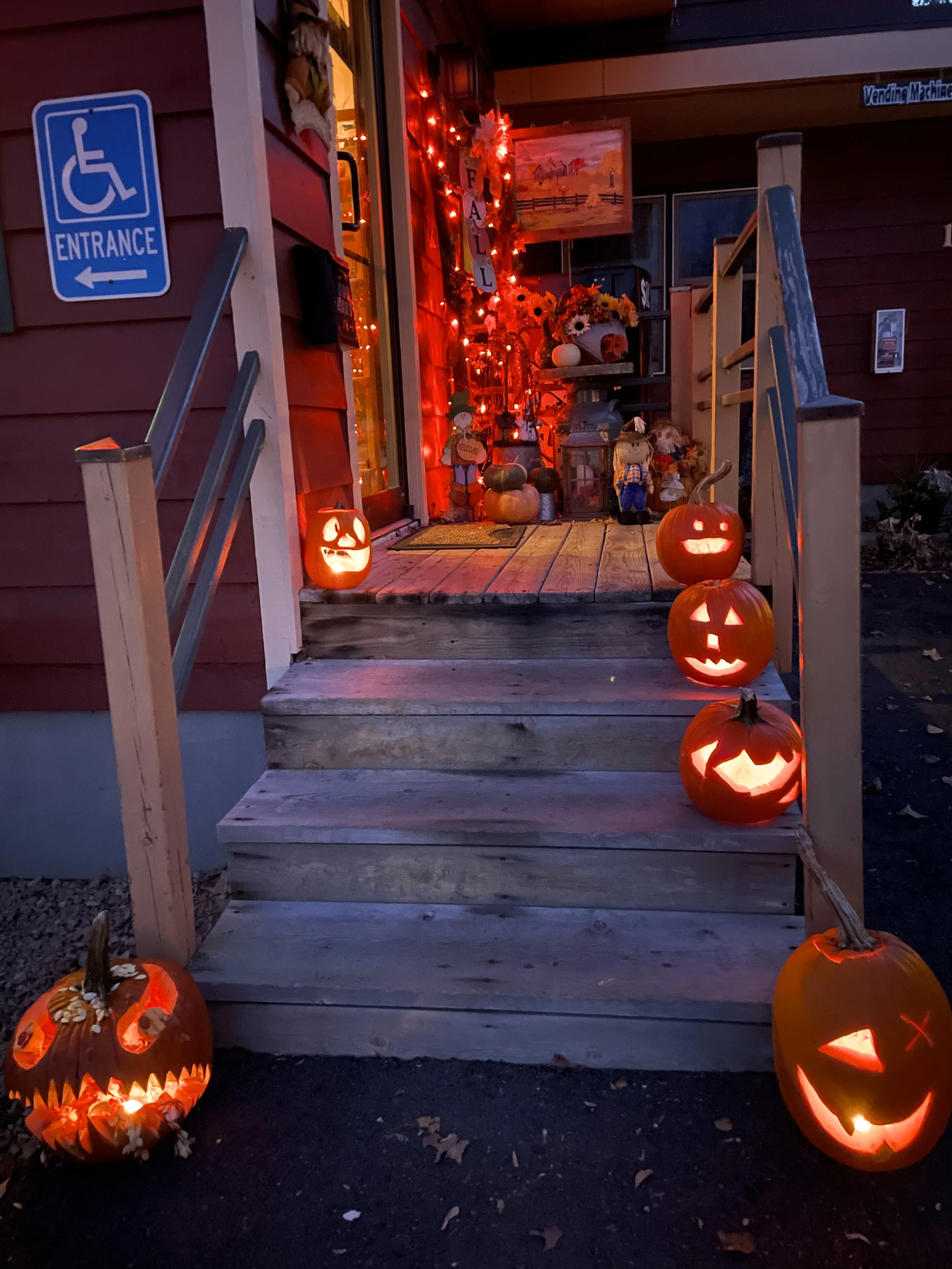 Decorated house entrance with pumpkins carved as jack-o'-lanterns, lit from inside, and Halloween decorations including scarecrows, flowers, and red string lights, with a porch staircase leading up to the door.