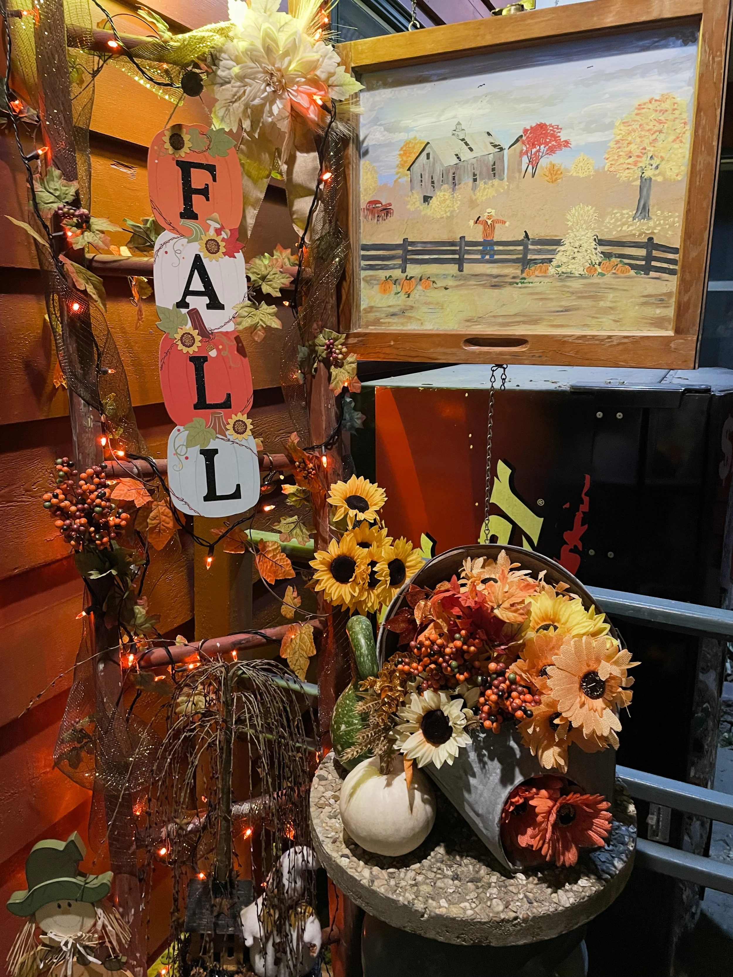 Autumn-themed porch decor with a vertical sign spelling 'FALL' surrounded by fall leaves and small fairy lights. There is a small pumpkin with flowers, a cone-shaped flower arrangement, and a colorful landscape painting of a farm scene with a person,