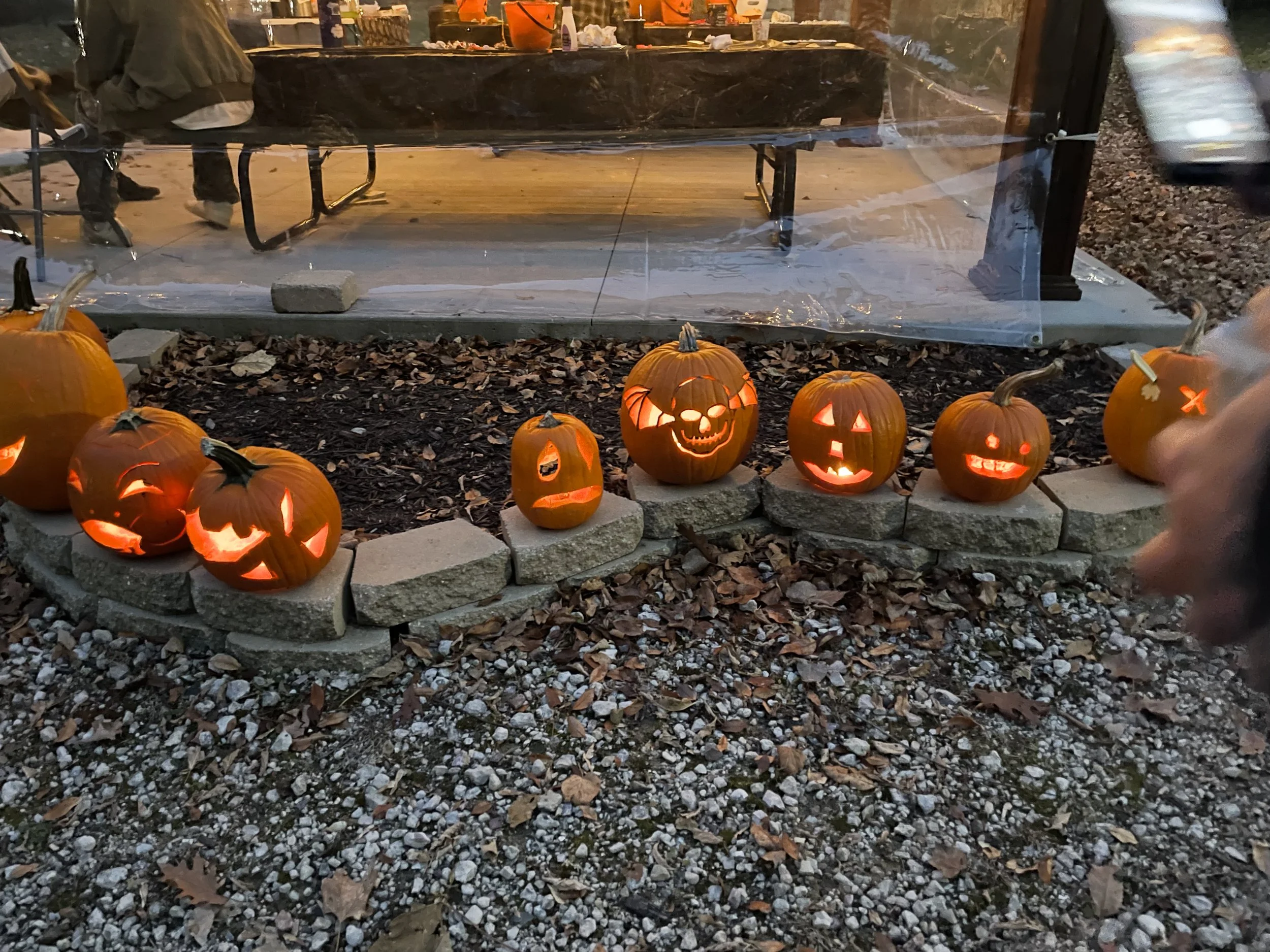 Line of illuminated carved Halloween pumpkins on a stone border outside, with a transparent enclosure and a table with people inside in the background.