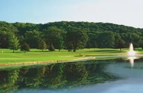 A scenic landscape featuring a calm lake with a fountain, surrounded by lush green grass and trees, with a hill in the background under a clear blue sky.