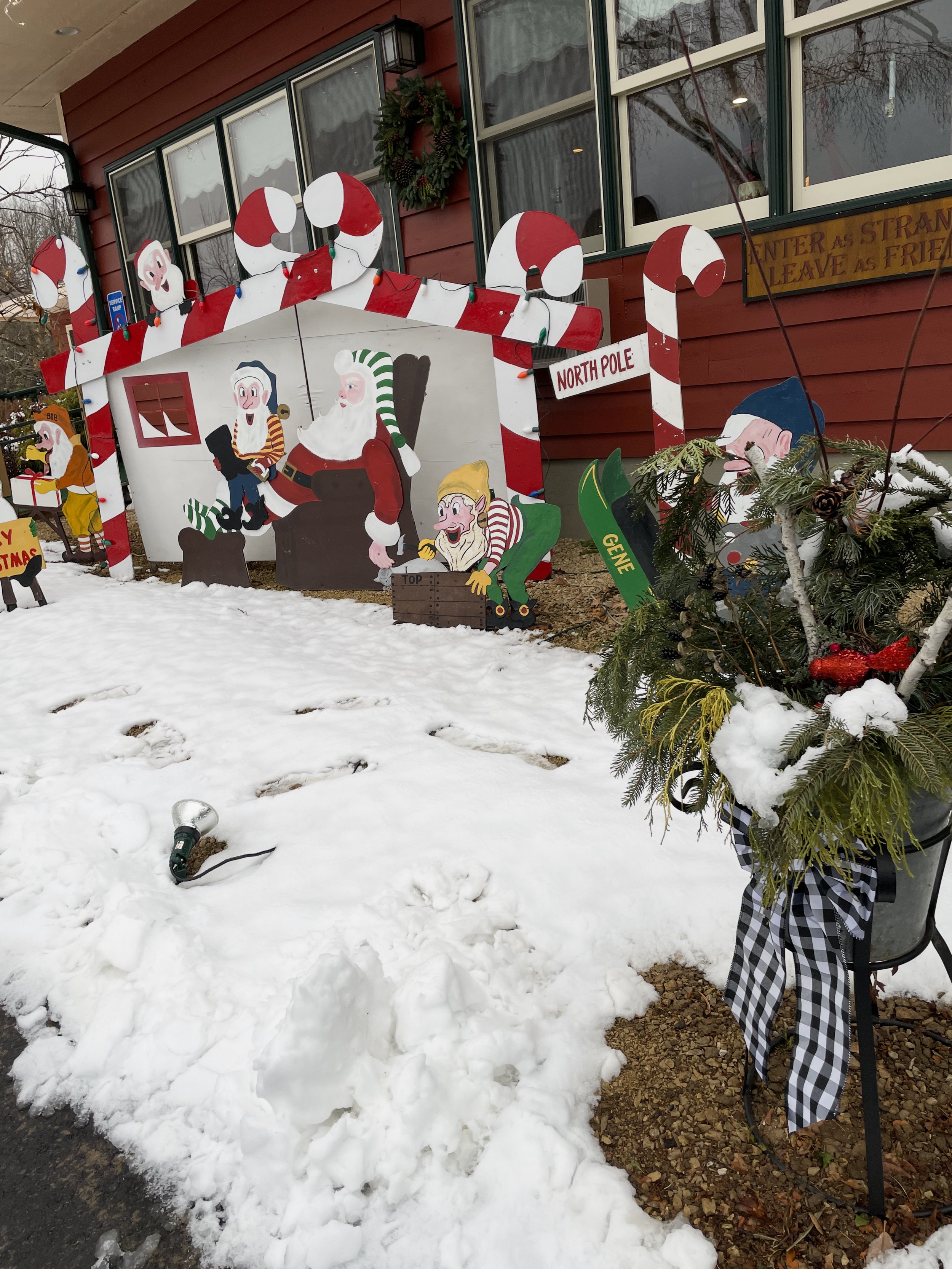 Decorative Christmas display featuring cartoon-like Santa Claus, elves, and candy cane props outside a building with snow on the ground.