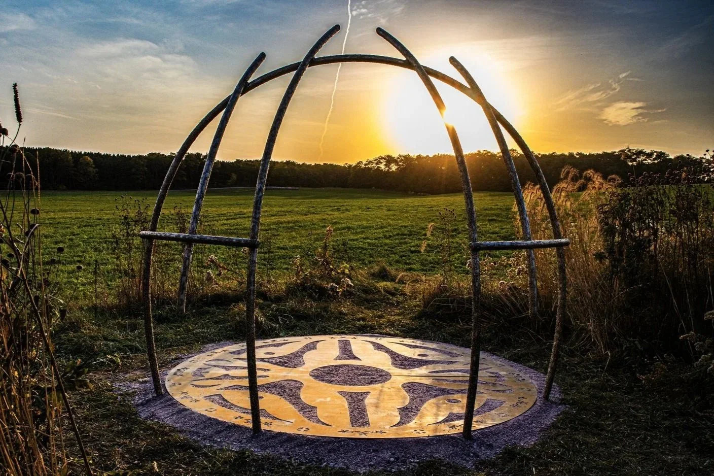 An outdoor sculpture resembling a clock face on the ground, with metal arches above, in a grassy field at sunset.
