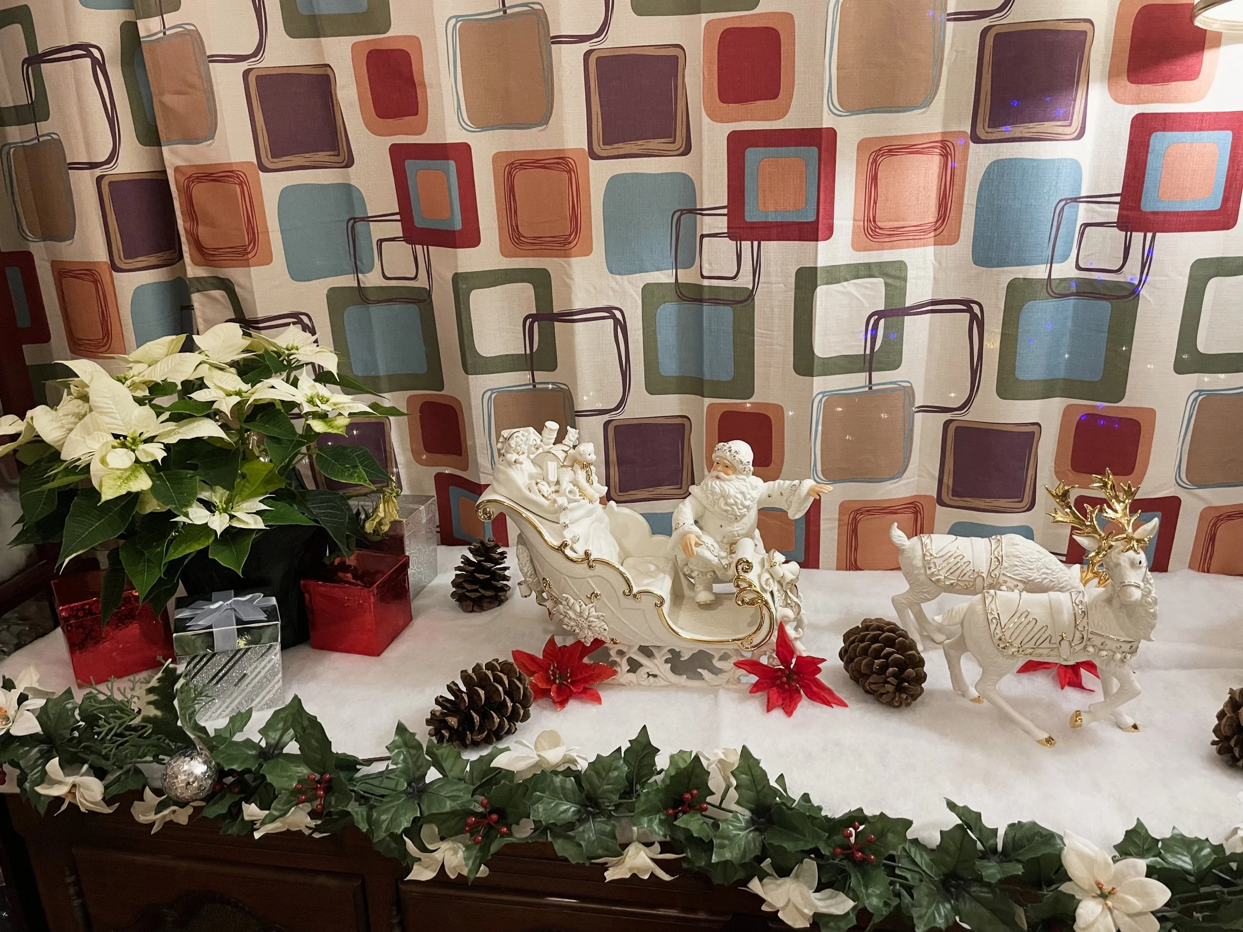 Christmas display with Santa Claus figurine in a sleigh, reindeer figures, poinsettia flowers, pinecones, and a poinsettia plant, all on a table decorated with holly and white flowers against a colorful square-patterned curtain background.