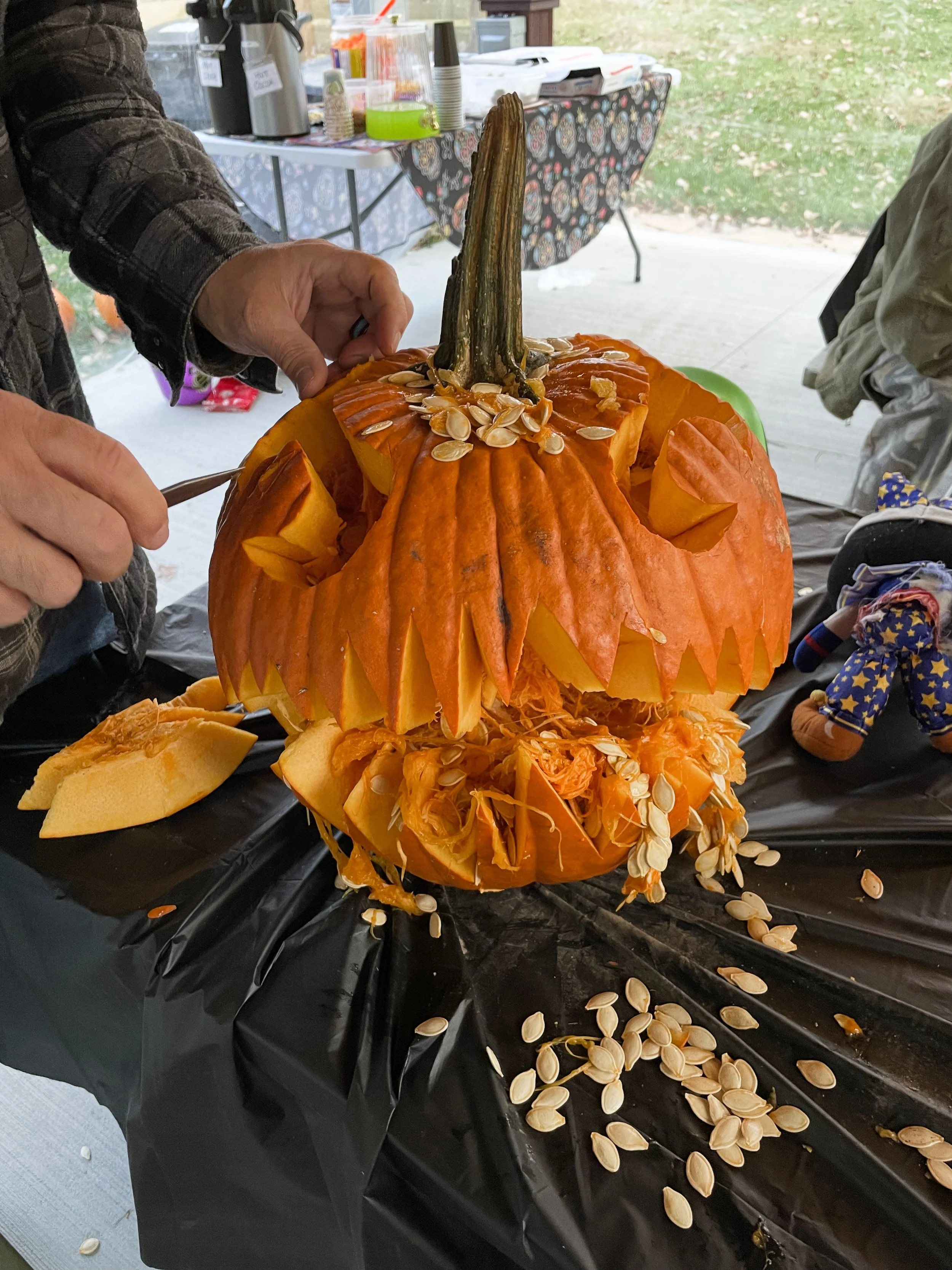 A carved pumpkin with a jagged mouth and eyes is being scooped out, with pumpkin seeds spilling onto a black tablecloth. The top of the pumpkin is removed, revealing the insides and seeds, and someone is working on it with a spoon.
