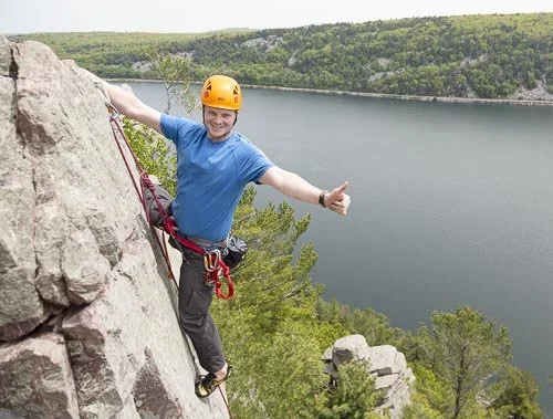 Person wearing a helmet rock climbing on a steep cliffside near a large body of water, giving a thumbs up.