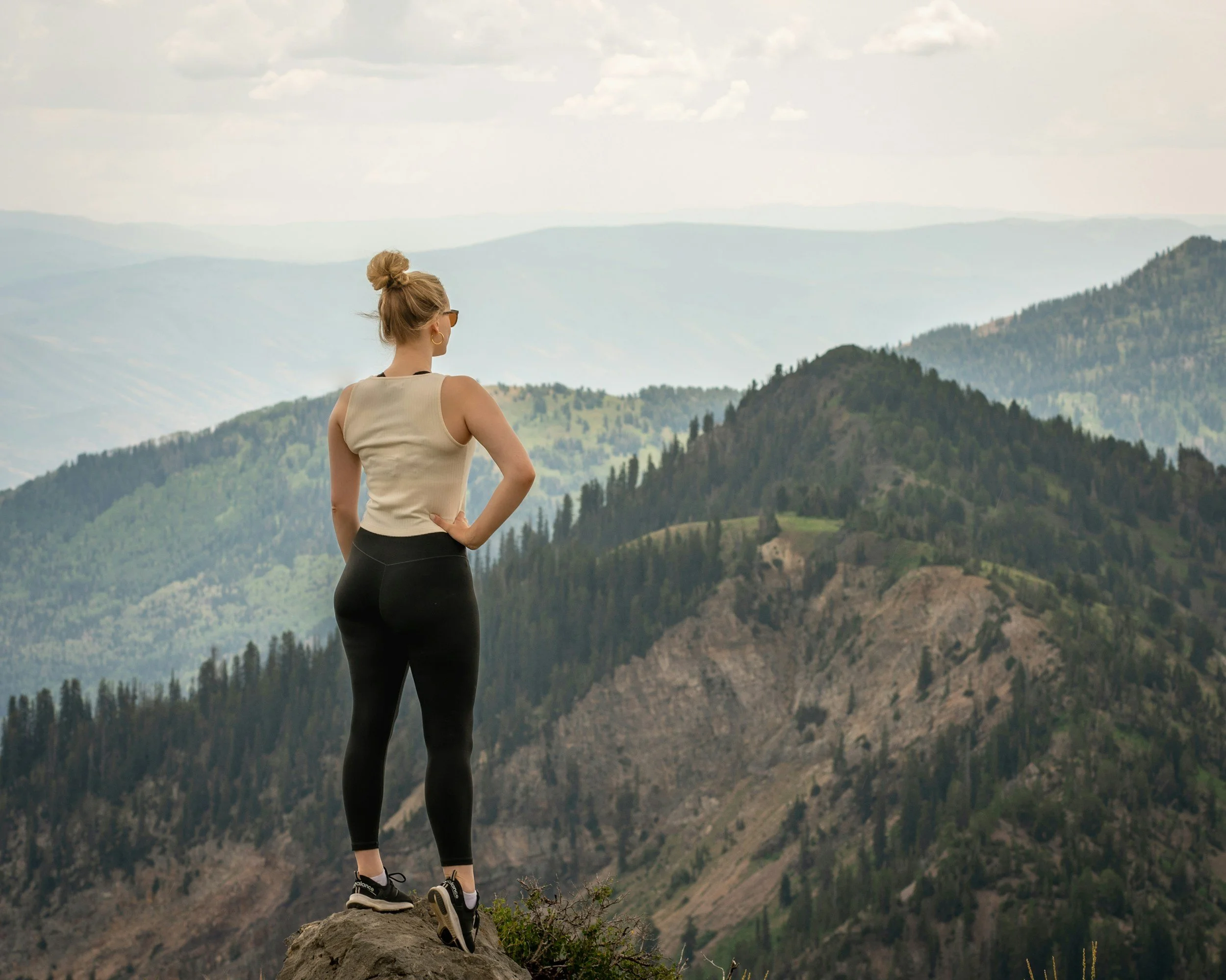 A woman stands on a rock overlooking green mountains and valleys, with her back to the camera, wearing a beige tank top, black leggings, and sneakers, while her hair is in a bun.