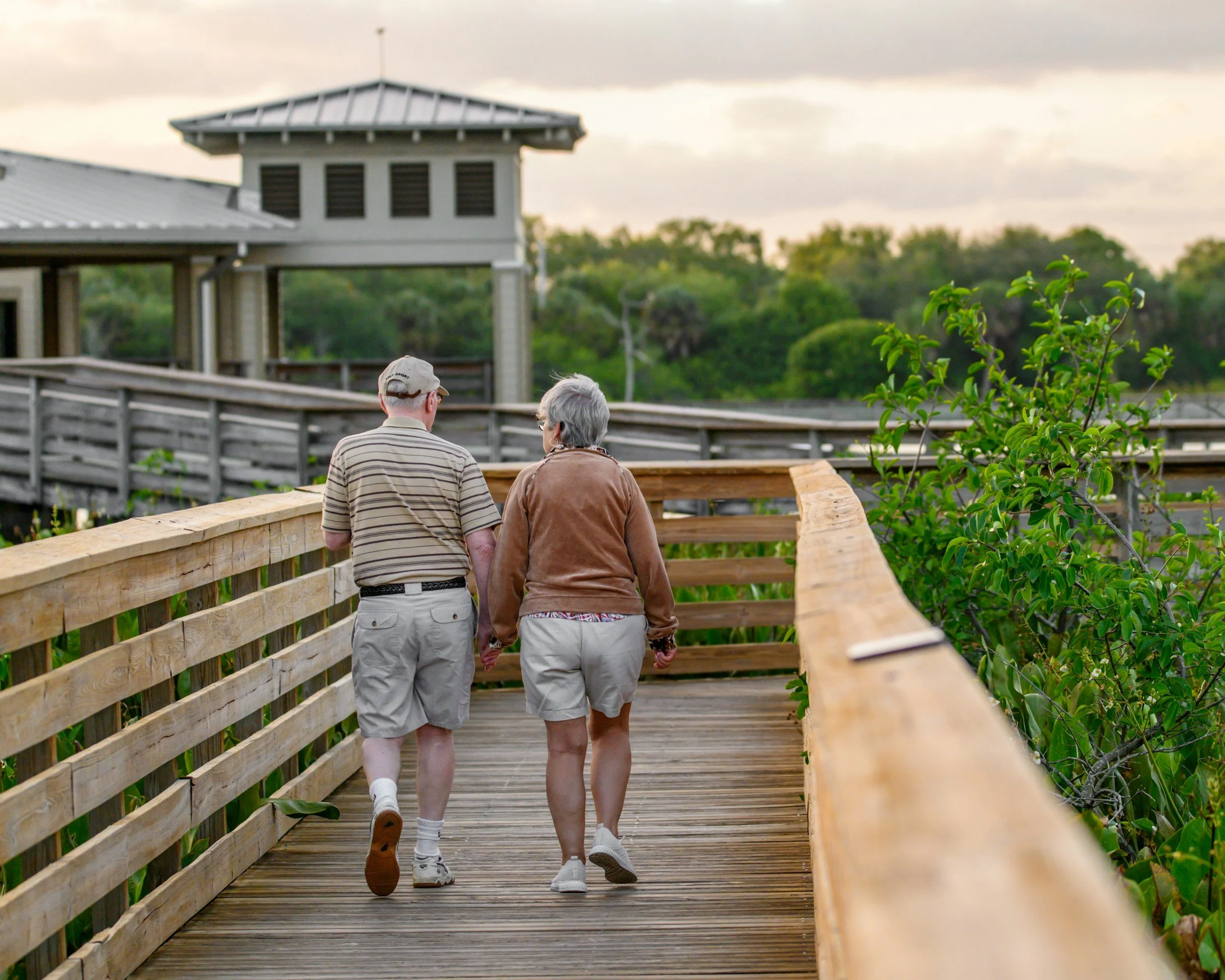 An elderly couple walking hand in hand on a wooden boardwalk in a park during daytime, surrounded by green trees and bushes.