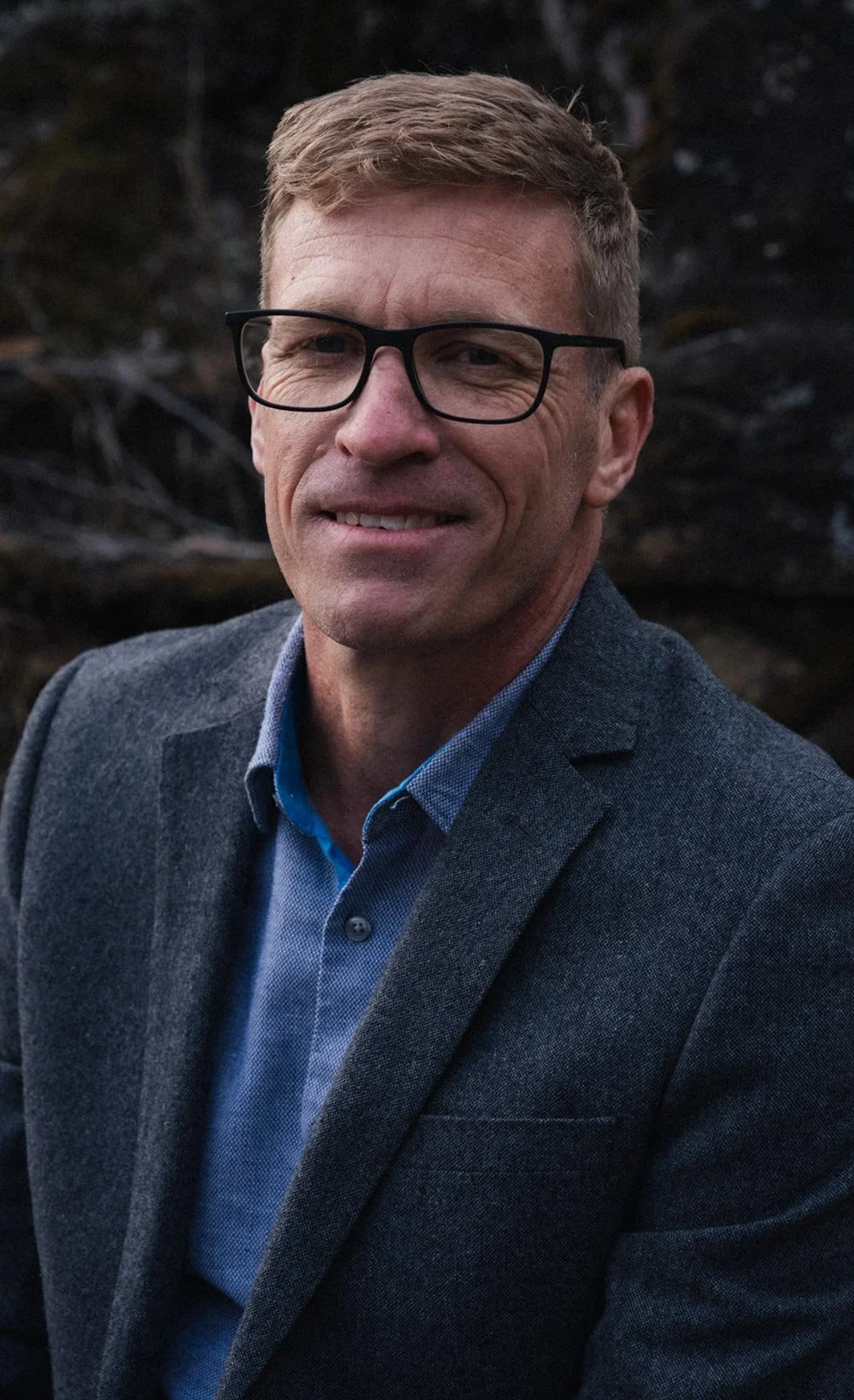 Dr David Spindler wearing a gray blazer and blue shirt, smiling outdoors with dark trees in the background.