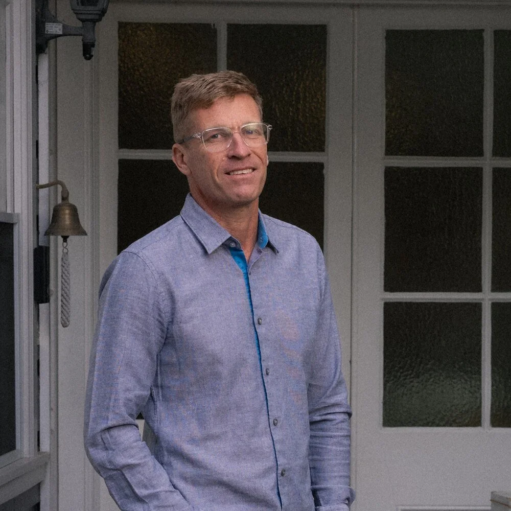 David Spindler, a man with glasses and light brown hair standing indoors near a window and door, wearing a blue button-up shirt.