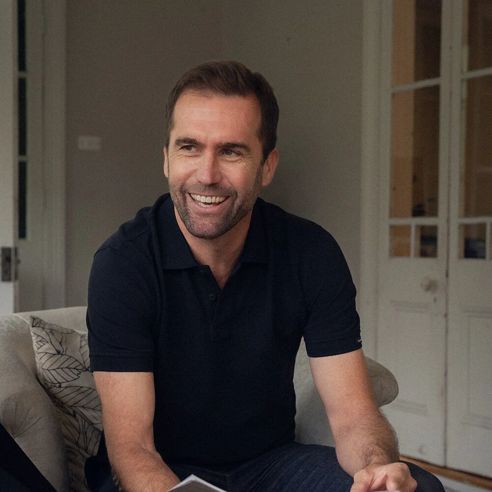 Brad McGee, a smiling man with dark hair and a beard, wearing a black polo shirt, sitting on a beige couch in a living room.