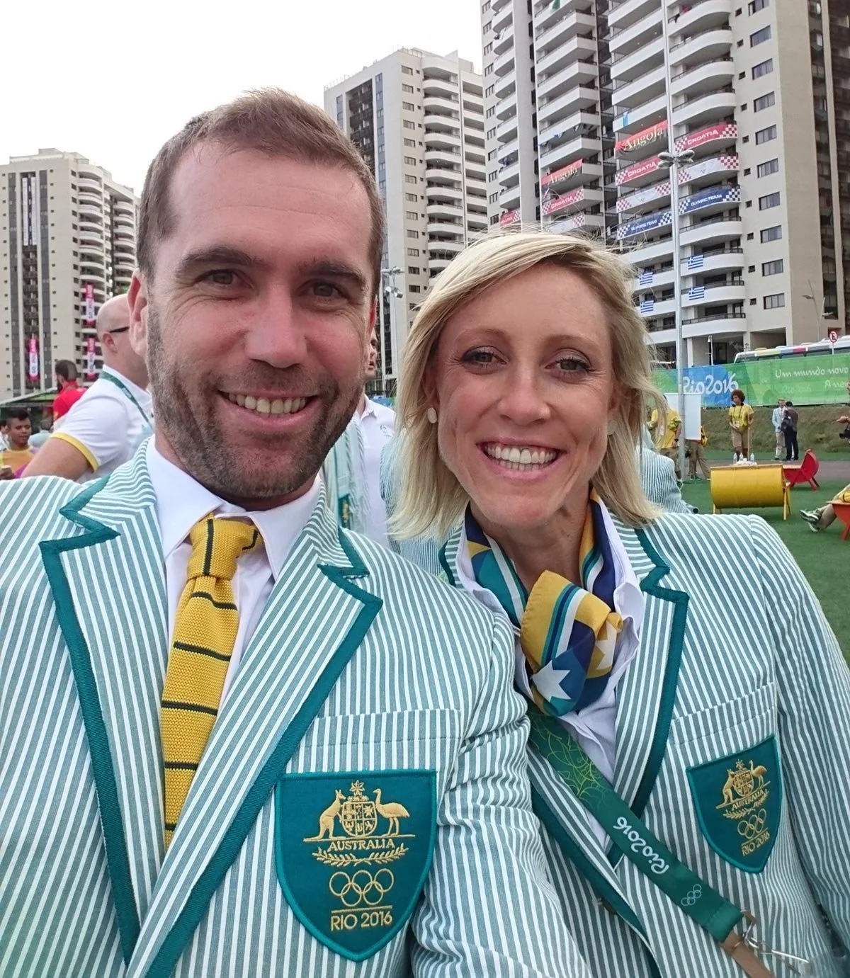 Australian Olympic team members Rachel Neylan and Brad McGee taking a selfie at the Rio 2016 Olympics, wearing teal and white striped blazers with Olympic badges, in an urban area with high-rise buildings and a Rio 2016 banner in the background.