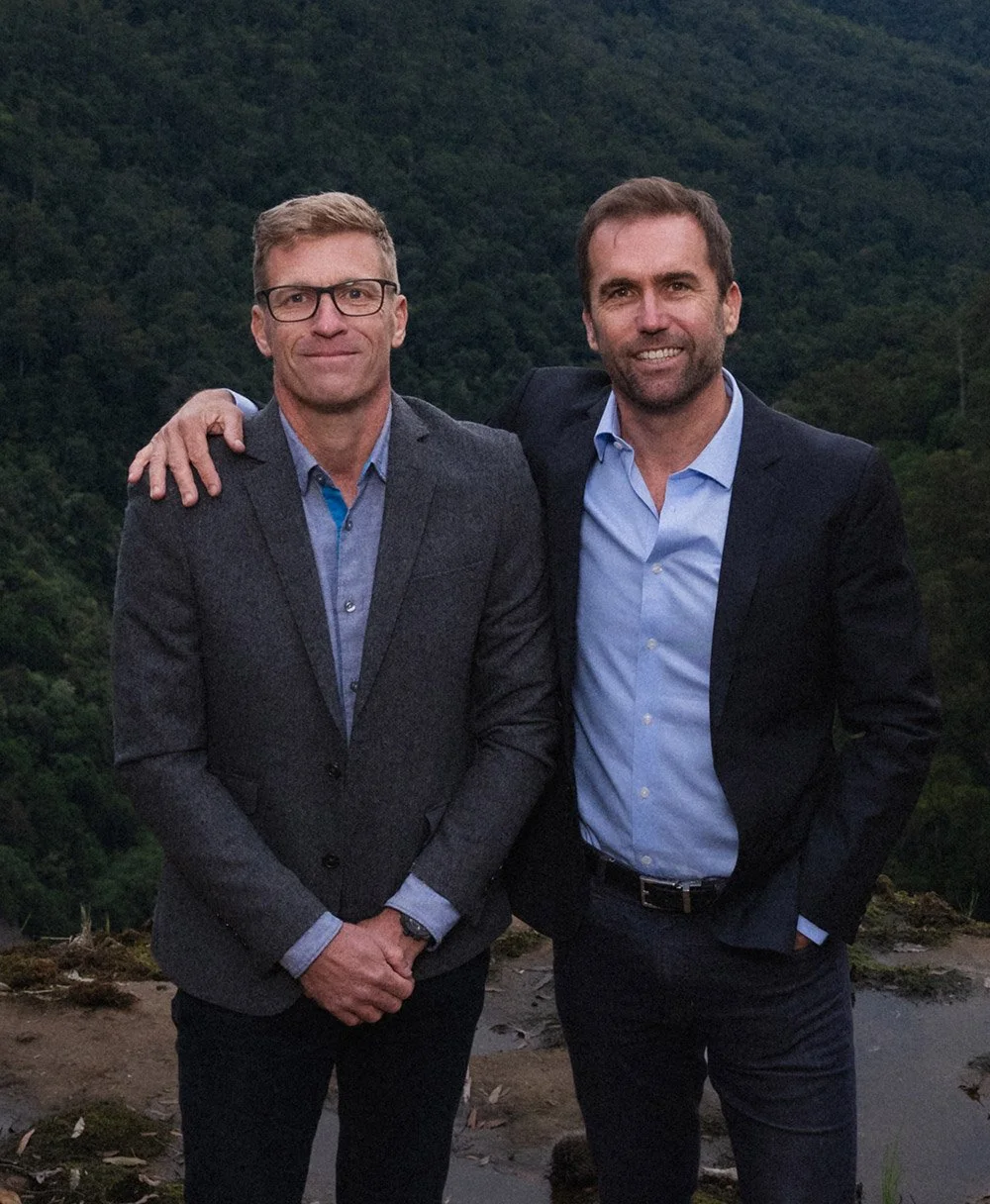 Brad McGee and David Spindler standing outdoors in front of a forested hillside, one with his arm around the other's shoulder, smiling.