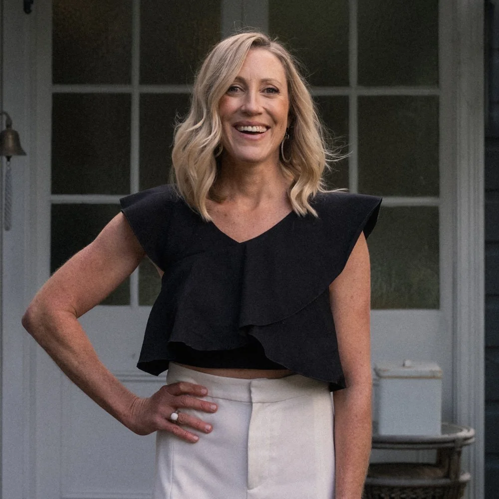 Rachel Neyland, a smiling woman with shoulder-length blonde hair, wearing a black ruffle sleeveless top and light-coloured high-waisted pants, standing outdoors in front of a house with a door and a window.